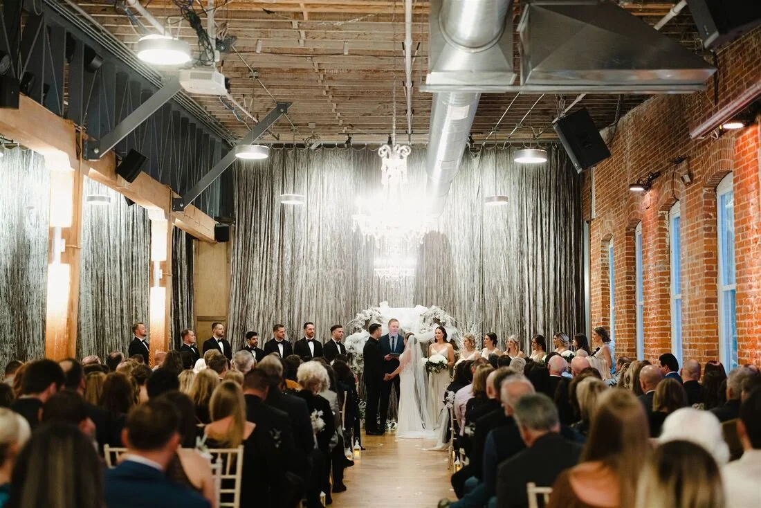 Beautiful indoor wedding ceremony with a bride, groom, officiant, bridesmaids, and groomsmen, in front of a decorative wall with drapes and a chandelier, filled with seated guests. Bright and lovely event space.