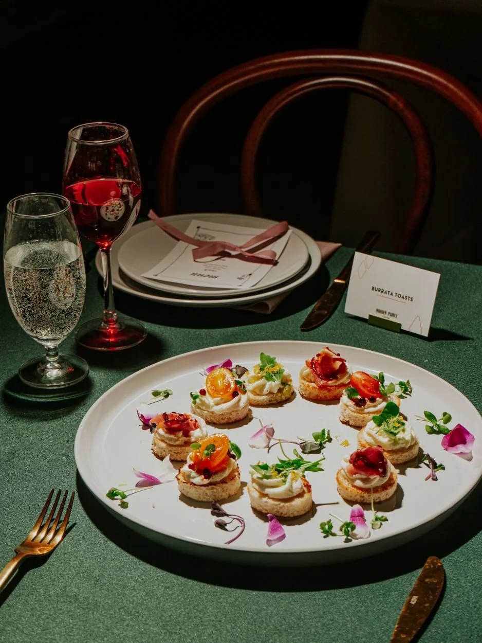 A dining table with a white plate of assorted canapé bites garnished with microgreens, flowers, and toppings like cherry tomatoes, cheese, and cured meats. Two glasses of sparkling water and a glass of red wine are next to the plate. There is a place setting with a menu, cutlery, and a small card labeled 'Burrata Toasts' on a green tablecloth.