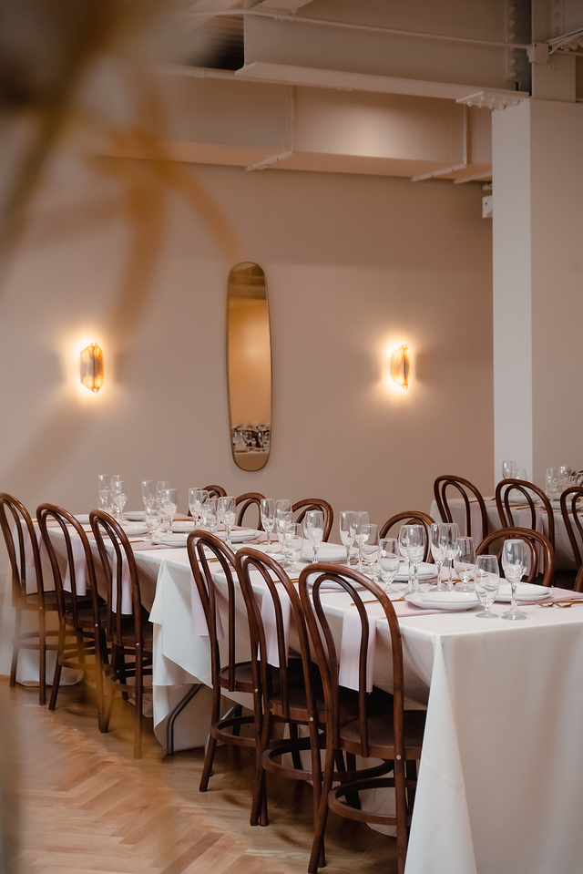 Long dining table with white tablecloth, set with glassware, napkins, and plates, surrounded by wooden chairs in a decorated restaurant or banquet hall.