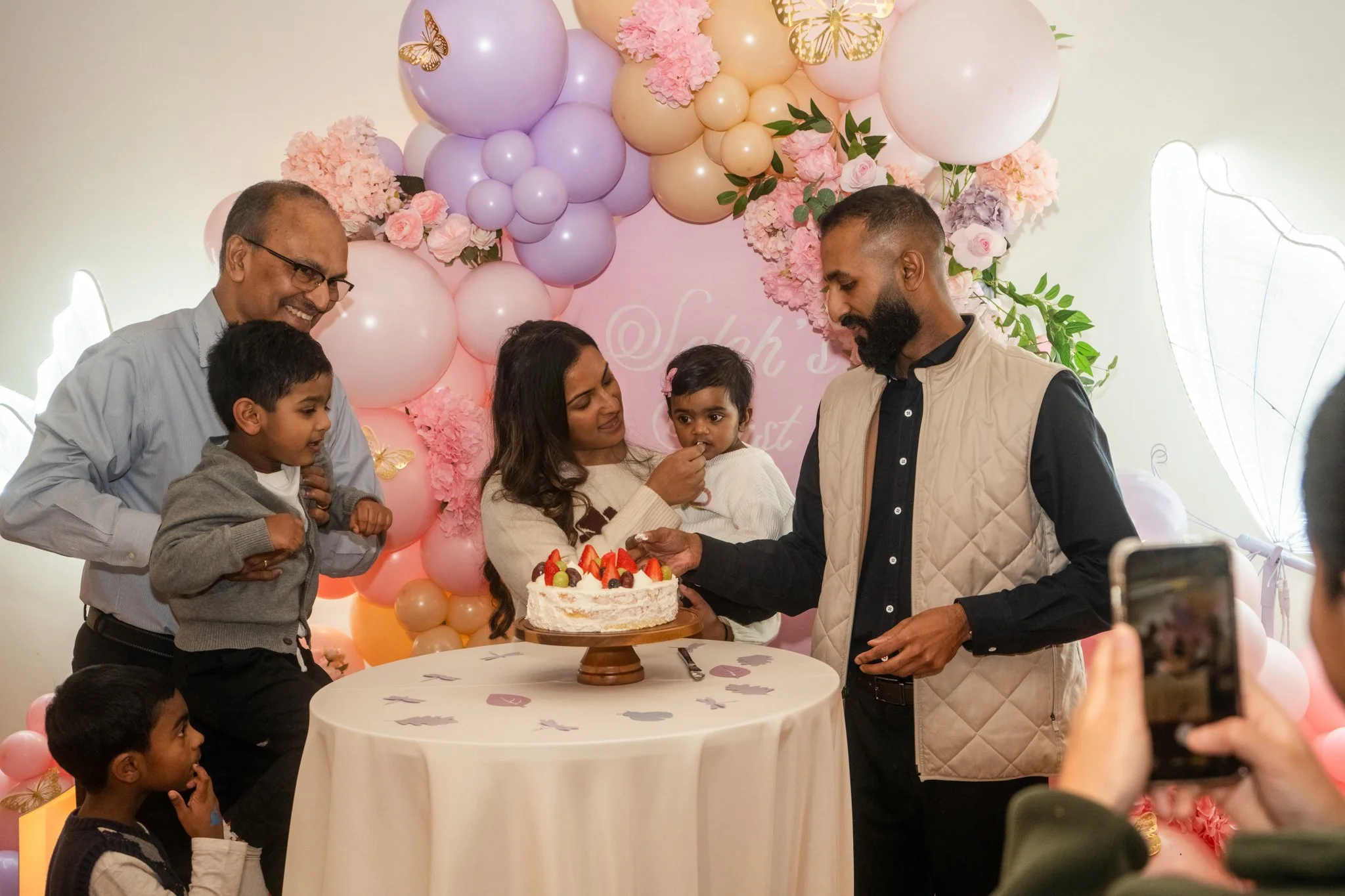 A family gathers around a birthday cake in front of a pink, purple, and peach balloon arch with pink flowers, celebrating a birthday. One woman holds a young child as another man holds a knife with a young girl in front of her. An older man and two other children also present, with a person taking a photo.