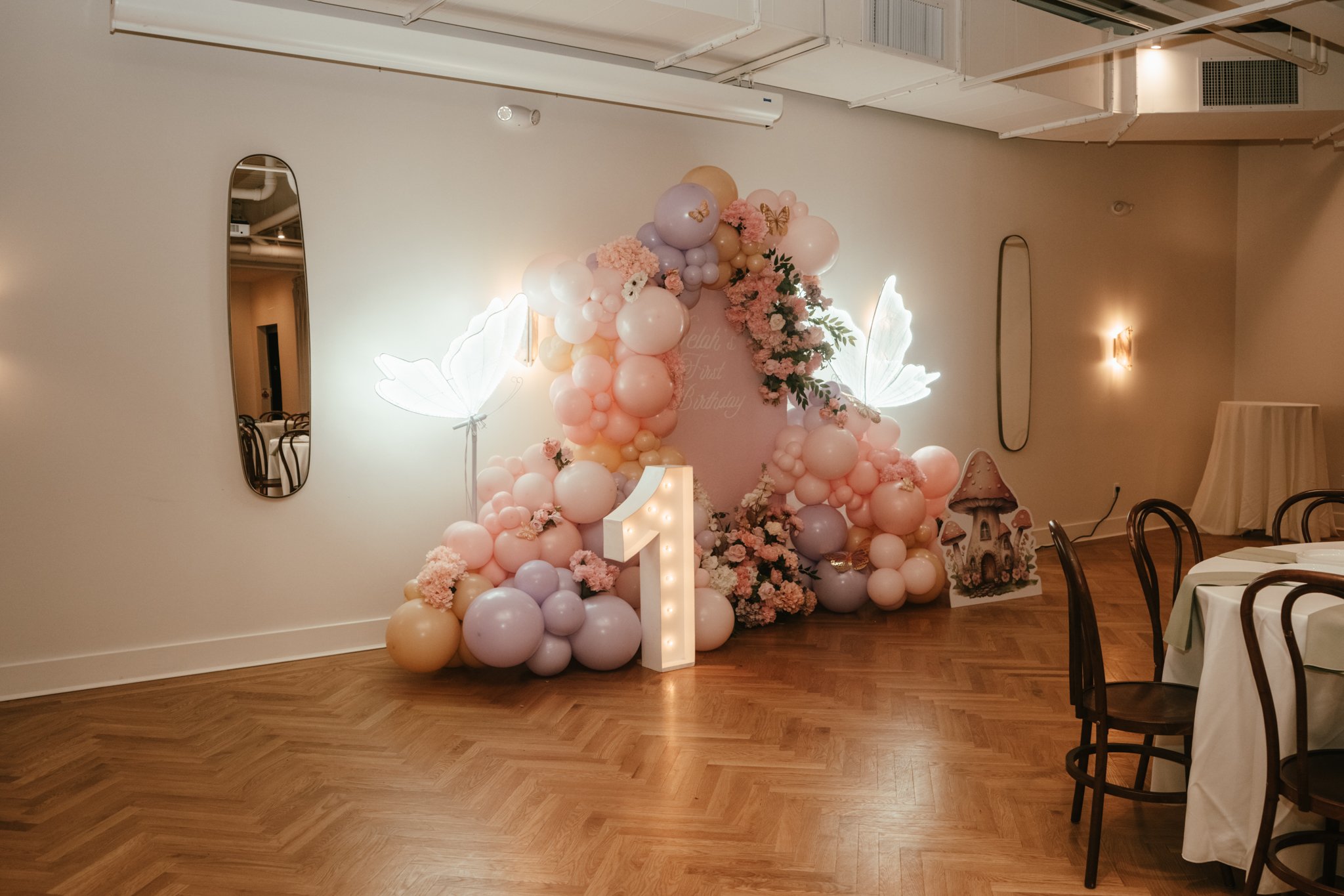 Decorative setup for a first birthday celebration with pastel pink, purple, and beige balloons arranged in an arch and clusters. Features illuminated butterfly wings, a large marquee number 1, and floral arrangements with pink and white flowers. The background includes a pink sign with faint text, whimsical mushroom decorations, and a simple, elegant interior with wooden flooring and mirrors.