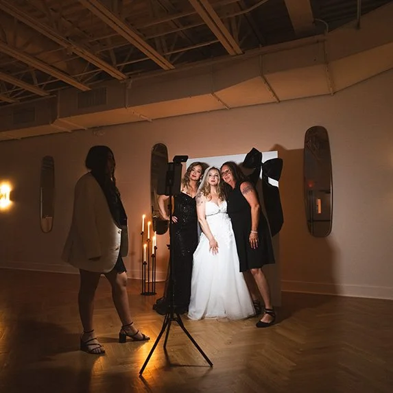 Four women, one in a white wedding dress, posing for a photo in a dimly lit indoor space with candles, mirrors, and a wooden floor.