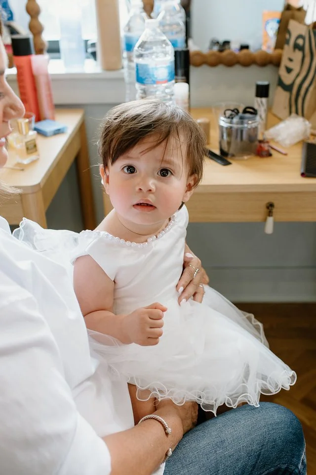 A young girl in a white dress with a pearl neckline sitting on an adult's lap indoors. Background includes a wooden table with water bottles, a backpack, and various items.