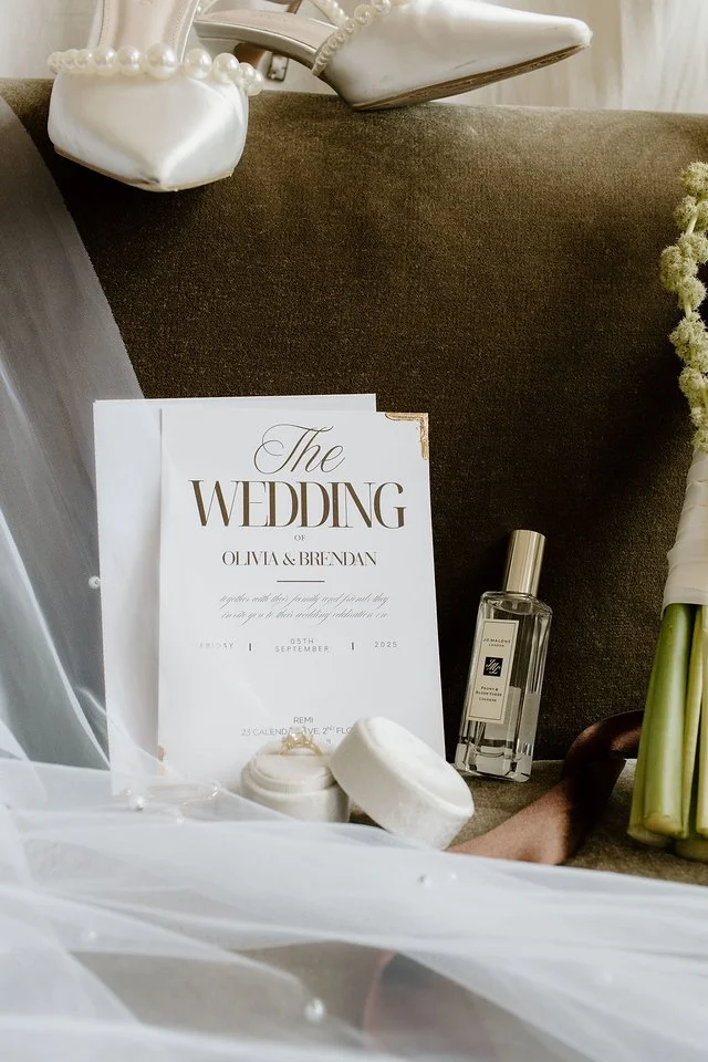Wedding invitation for Olivia & Brendan, dated September 5, 2025, placed on a brown surface, accompanied by white wedding accessories including a bouquet, perfume, and decorative items.