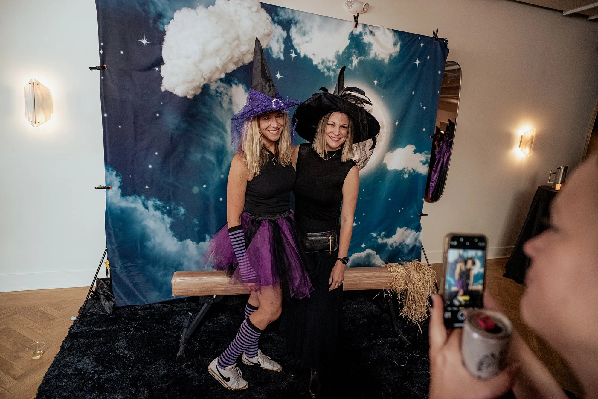 Two women dressed as witches with purple and black pointed hats, posing in front of a cloud and moon-themed backdrop at a costume party, with one woman taking a photo on her phone.
