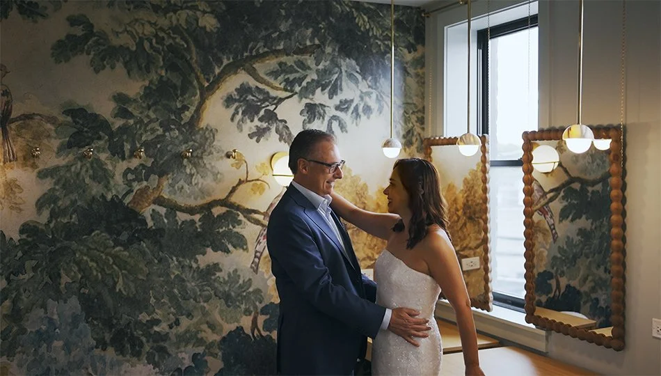 A man and a woman are dancing closely indoors, smiling at each other. The woman is in a white wedding dress, and the man in a dark suit. The room has a large window, wall-mounted lights, and artistic wallpaper with tree and branch illustrations.