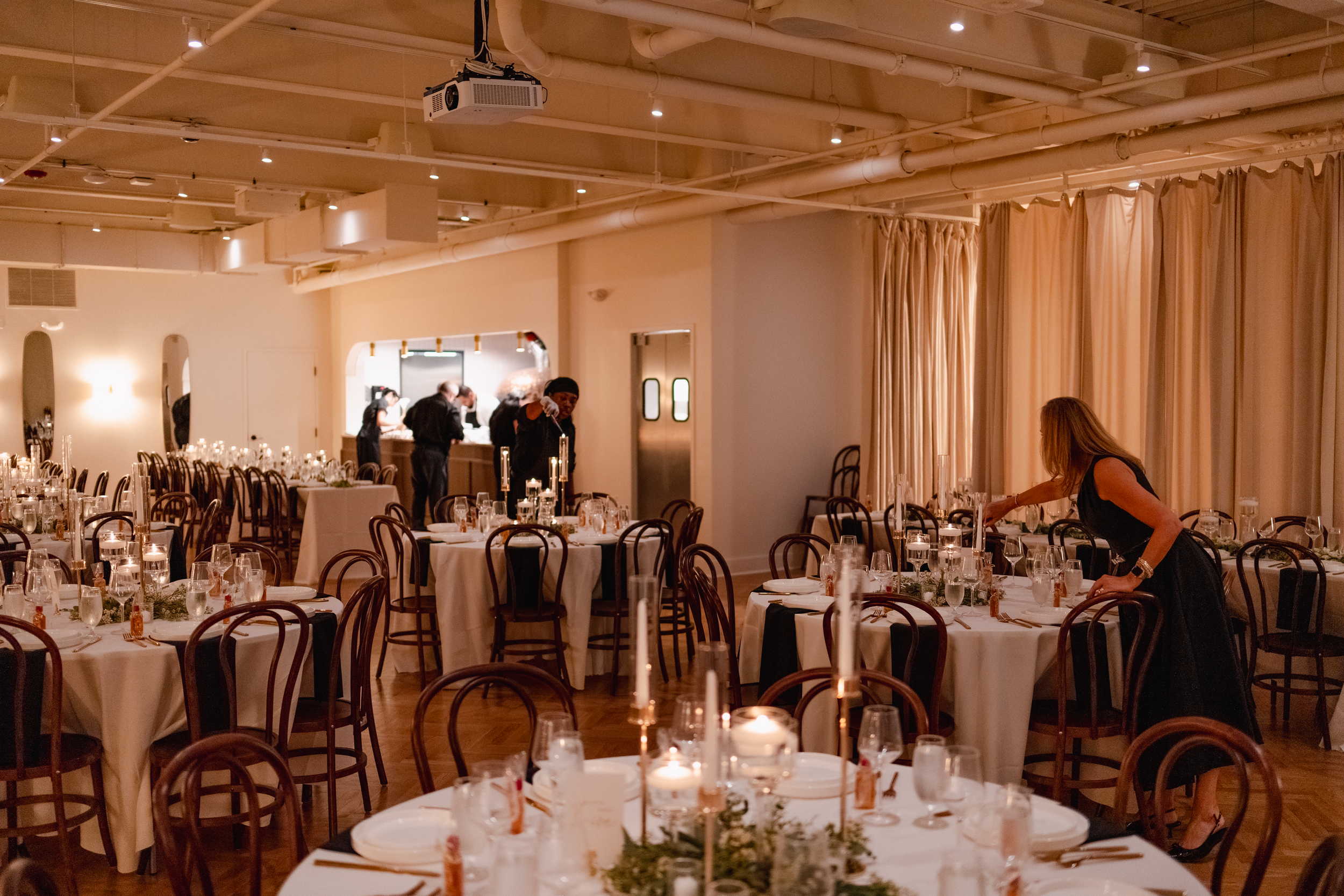 Elegant banquet hall with set tables, candles, and chairs, with people decorating and preparing for an event in the background, warm lighting, and cream-colored curtains.