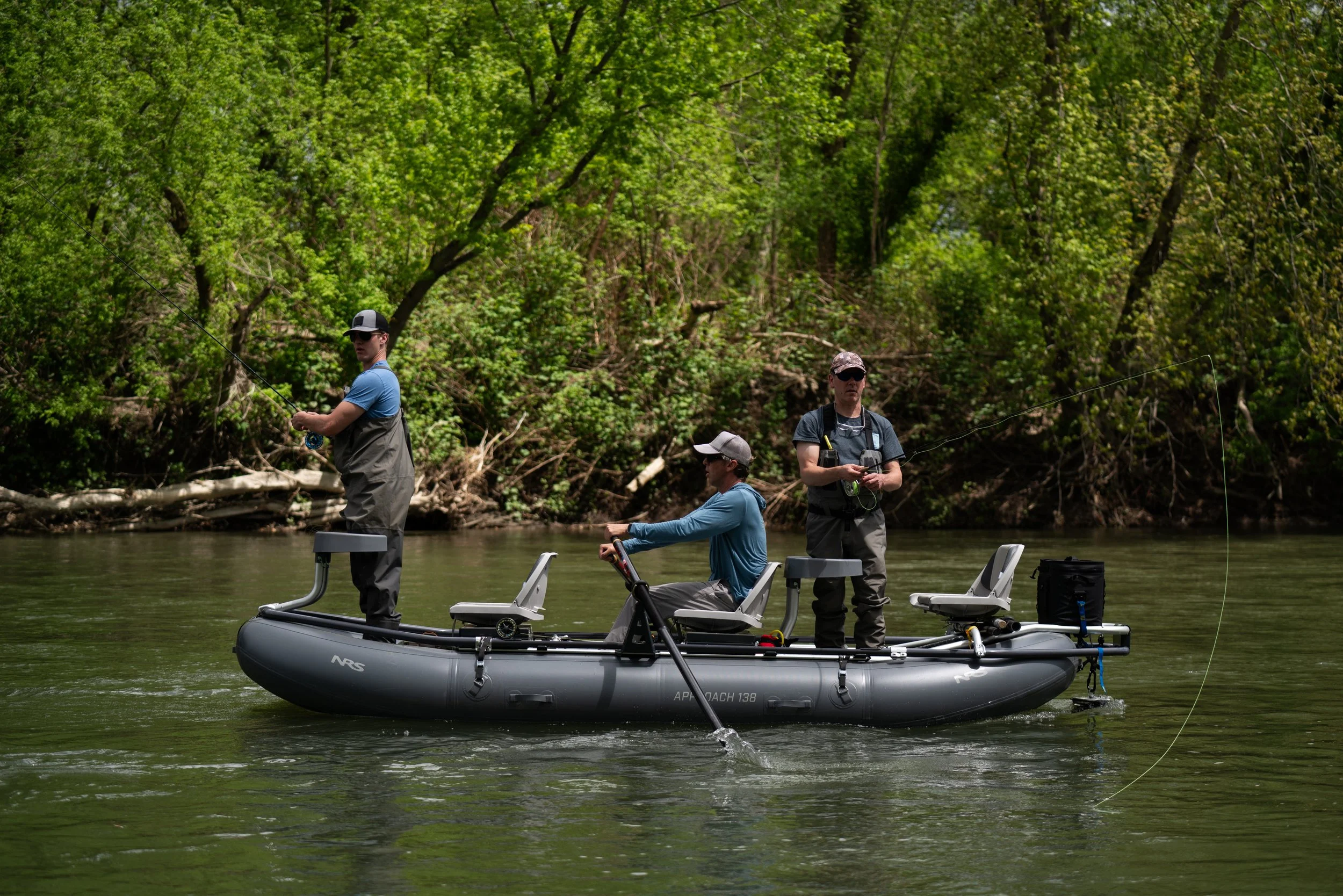 Three men fishing from an inflatable boat on a river, surrounded by green trees.
