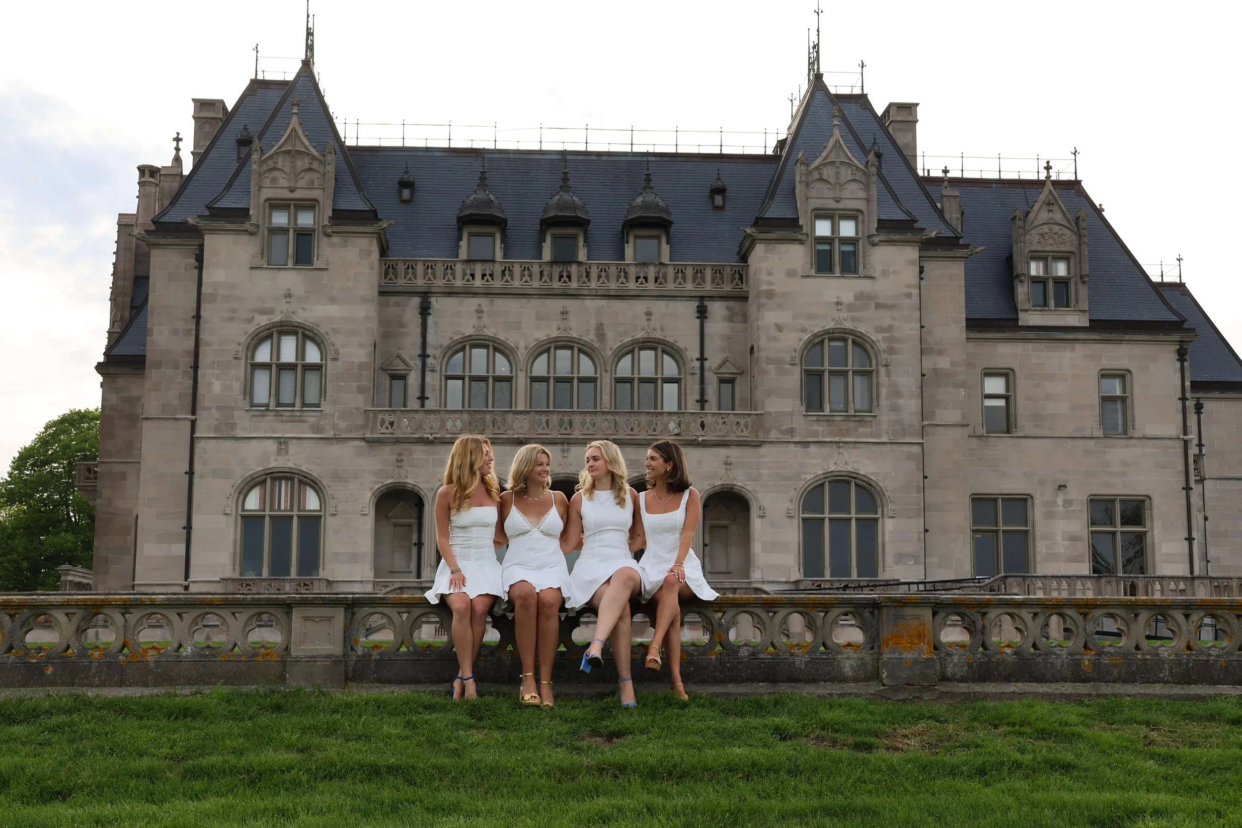 Four women in white dresses sitting on a stone fence in front of a large castle-like building with pointed roofs and multiple windows.