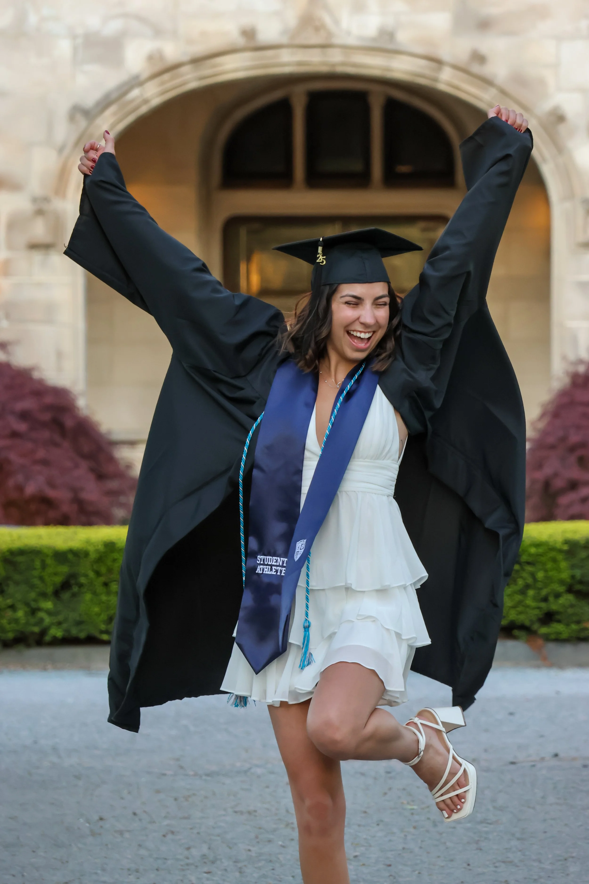 A woman in a graduation cap and gown celebrating outside a university building.