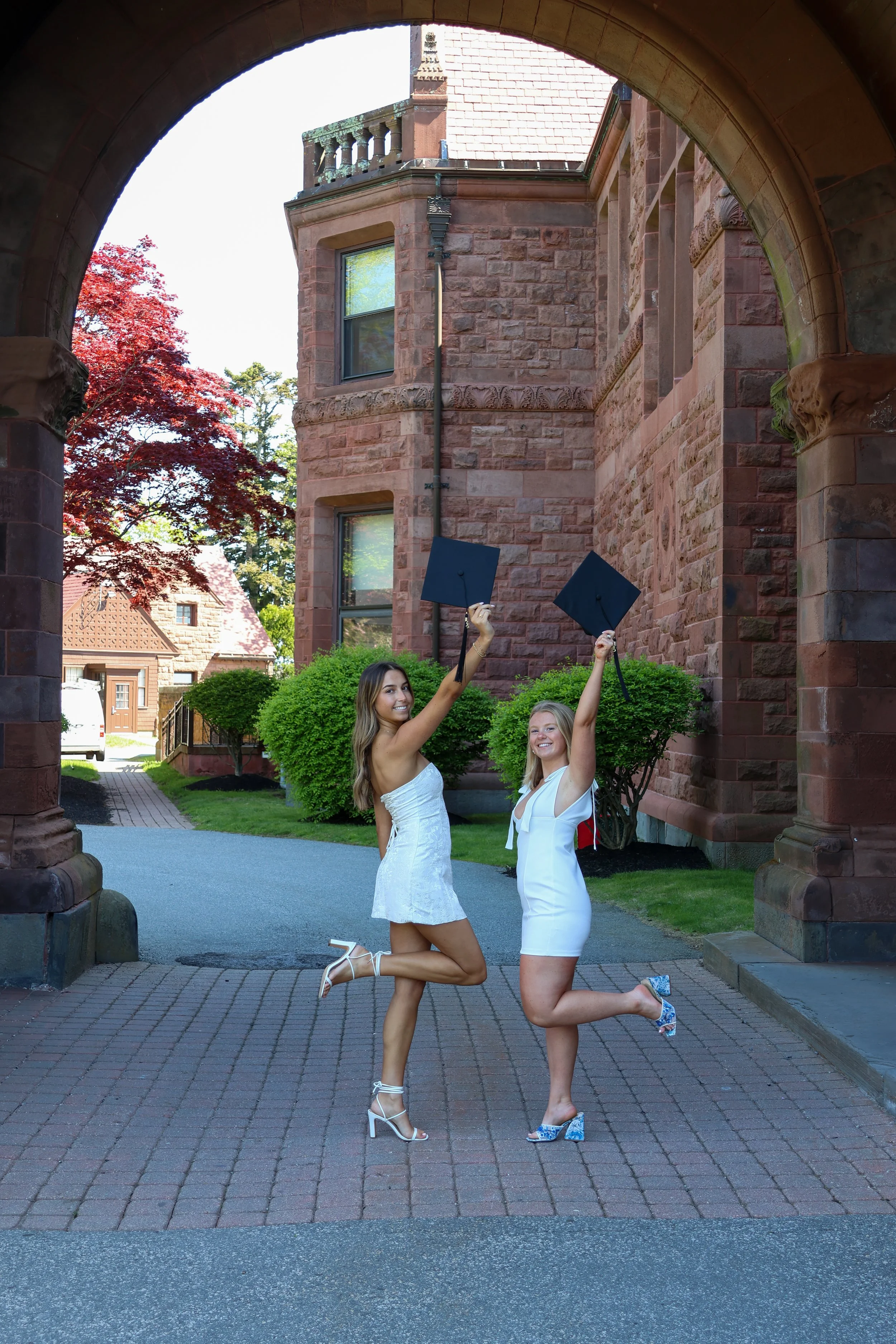 Two young women in white dresses celebrating graduation, holding up caps under a stone archway with a brick building and green trees in the background.