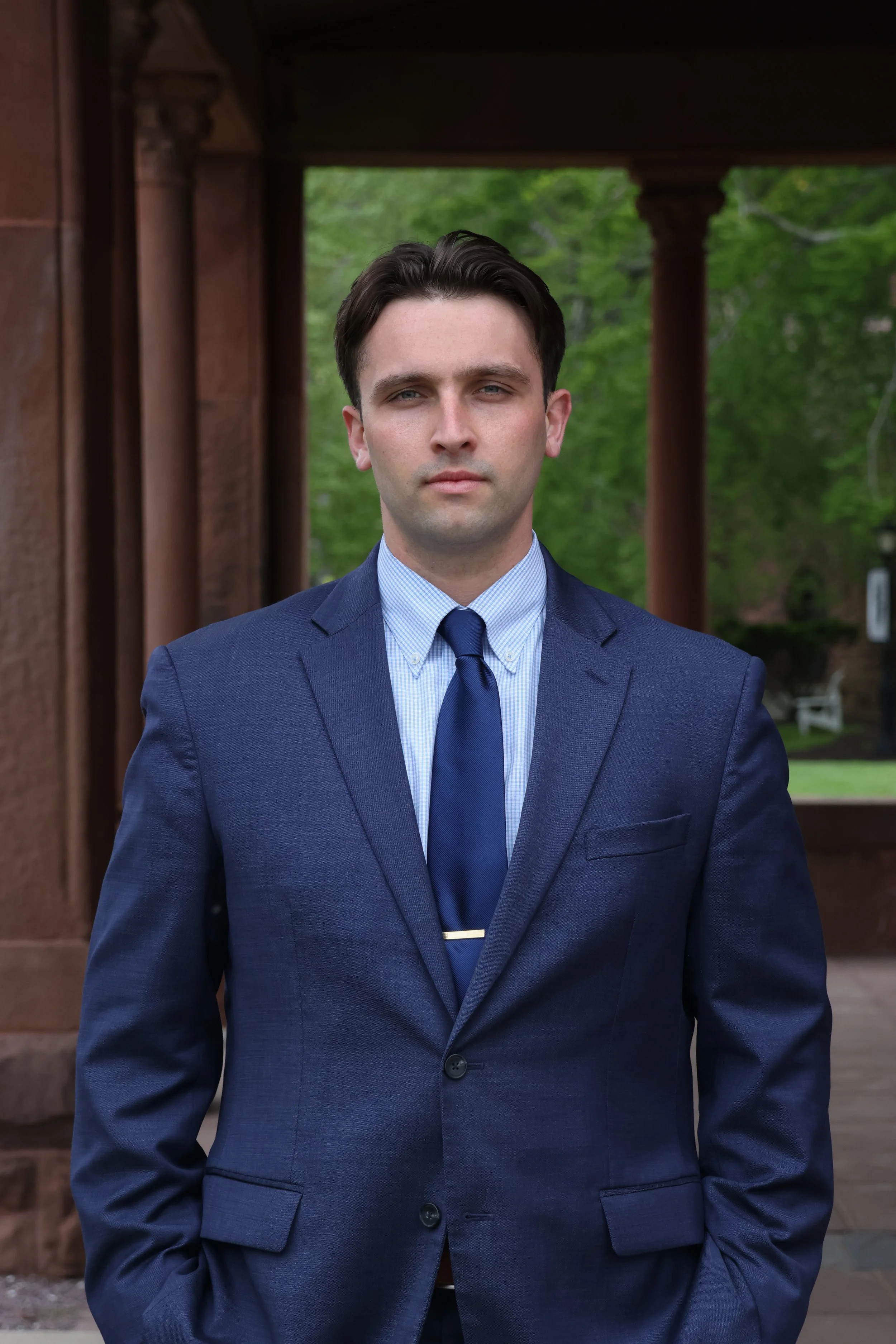 A young man in a navy blue suit and tie stands outdoors in front of a wooden structure with green trees in the background.
