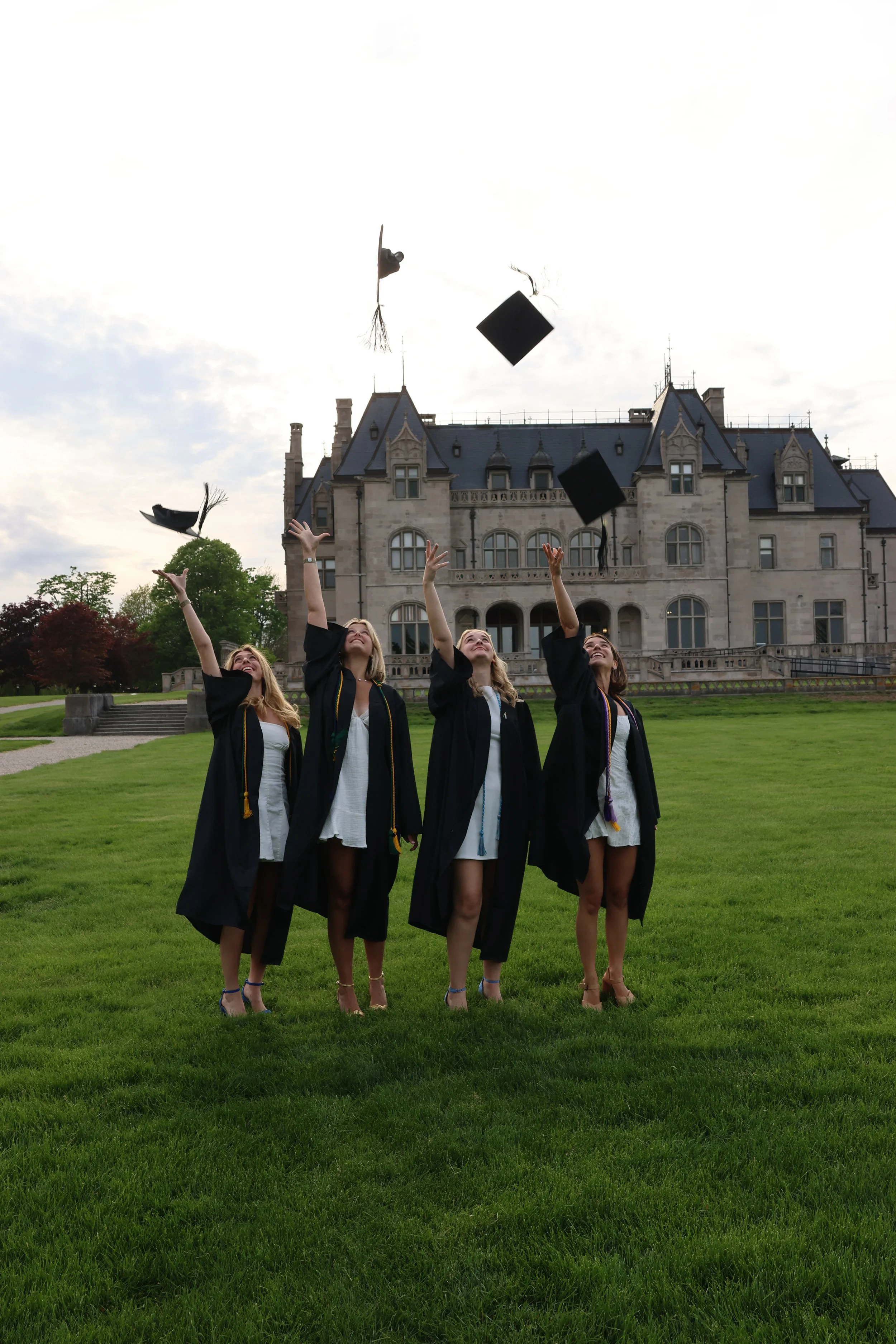 Four women in graduation gowns and caps celebrating outdoors in front of a large castle-like building, tossing caps into the air.