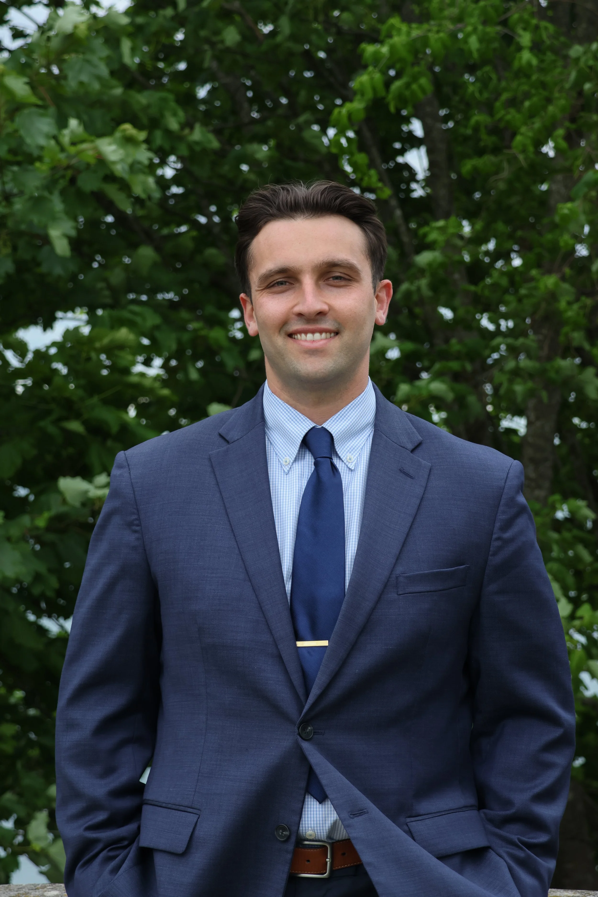 A young man in a navy blue suit, light blue dress shirt, and blue tie, smiling outdoors with green trees in the background.