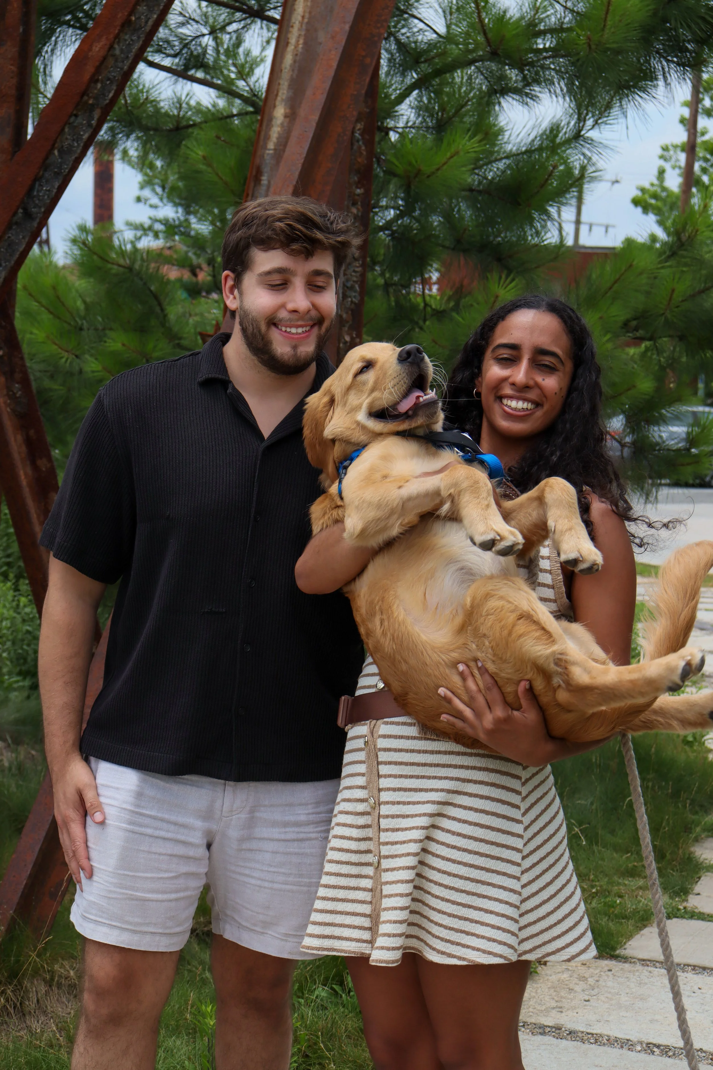 A young woman holding a golden retriever puppy while smiling, standing next to a young man outdoors with green trees and a sidewalk in the background.