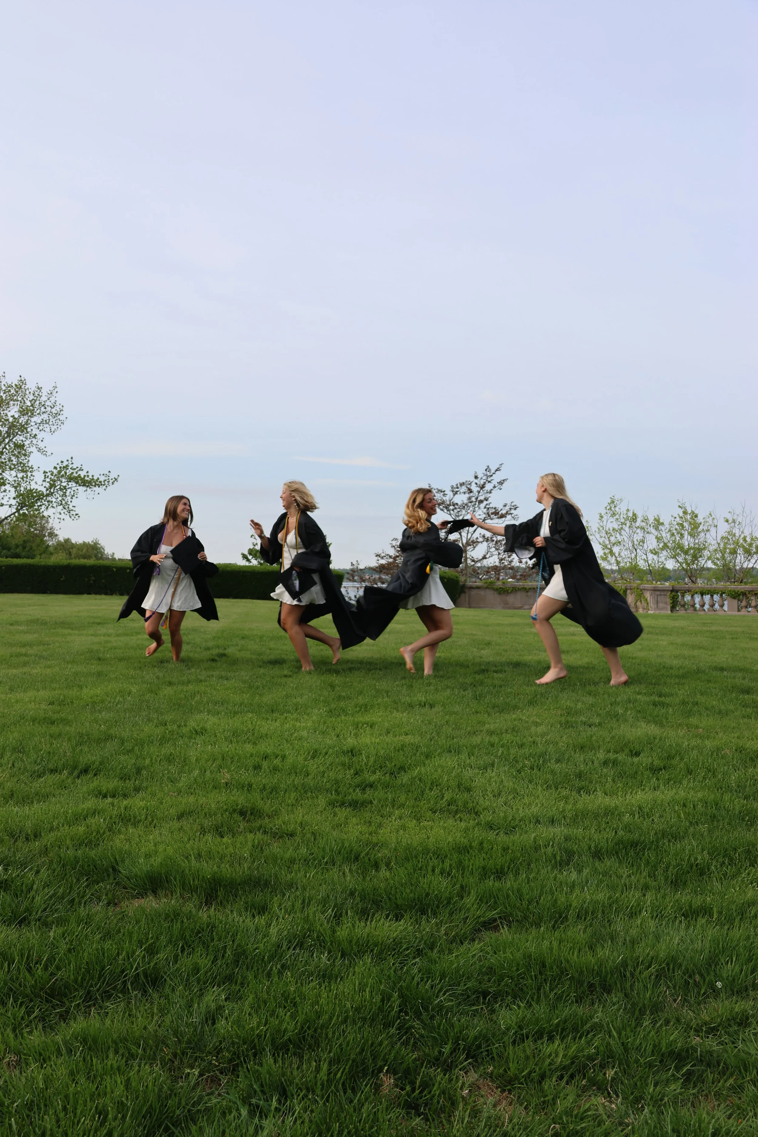 Four women in graduation gowns celebrating together on a grassy field.