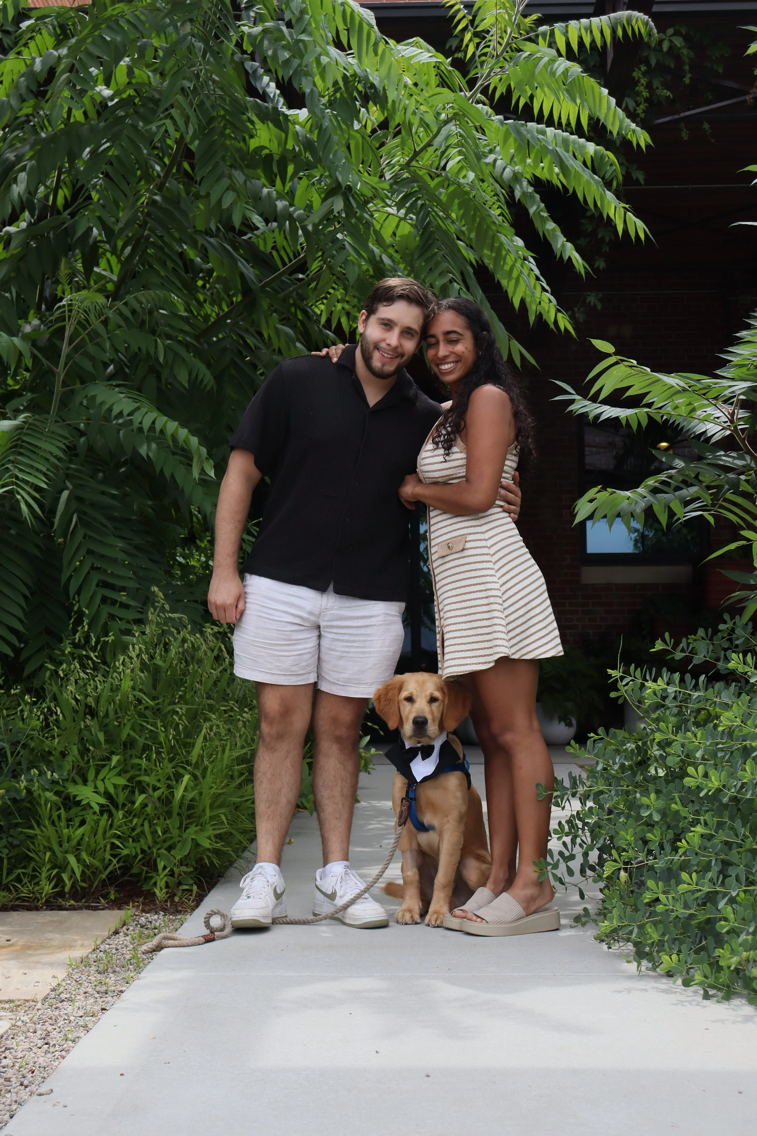 A smiling couple stands outside on a sidewalk with their golden retriever puppy dressed in a tuxedo costume. The man wears a black shirt and white shorts, and the woman wears a beige striped dress. The background features lush green plants and a buil