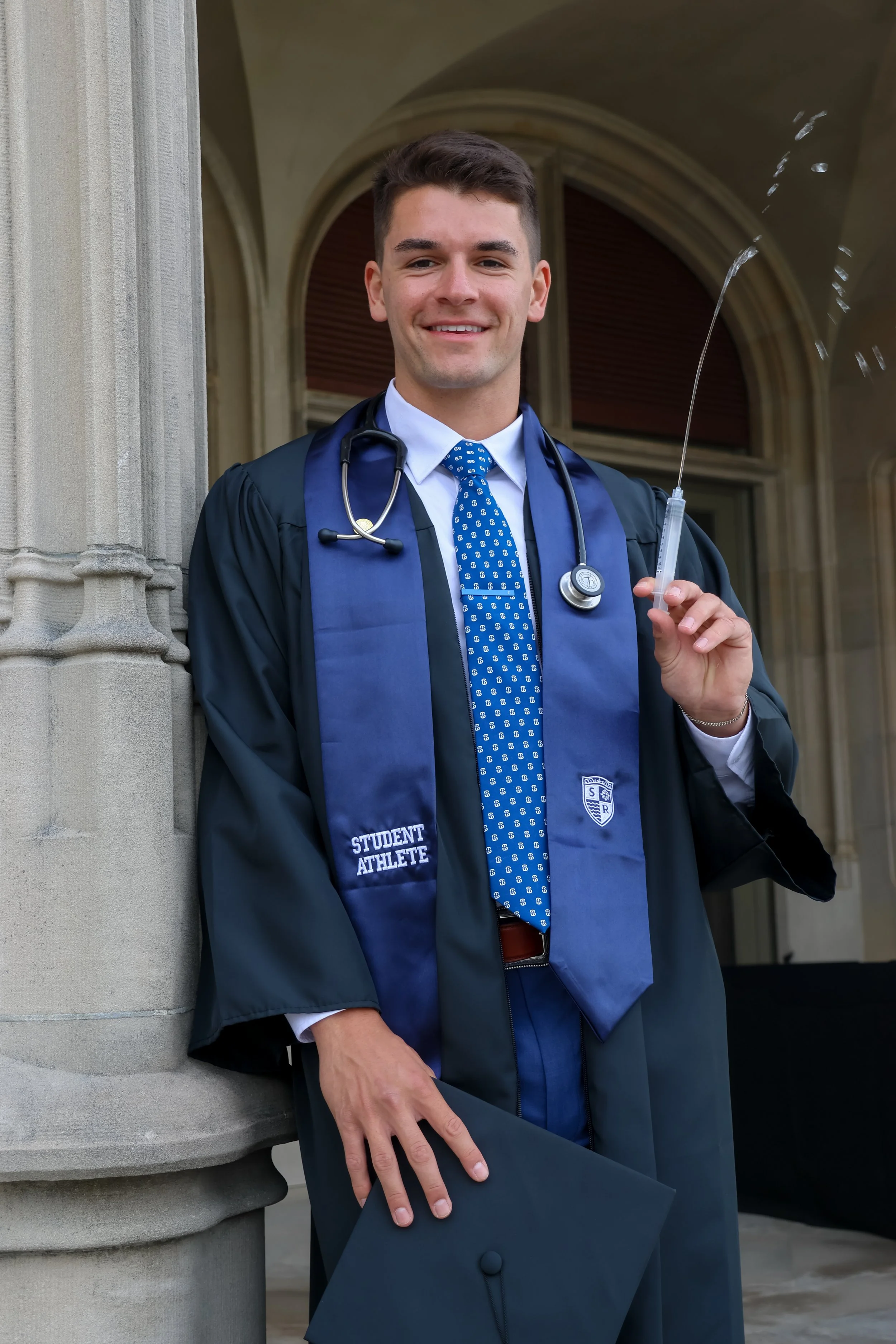 A young man in a graduation gown and cap holding a diploma, with a stethoscope around his neck, standing outdoors in front of an architectural building.