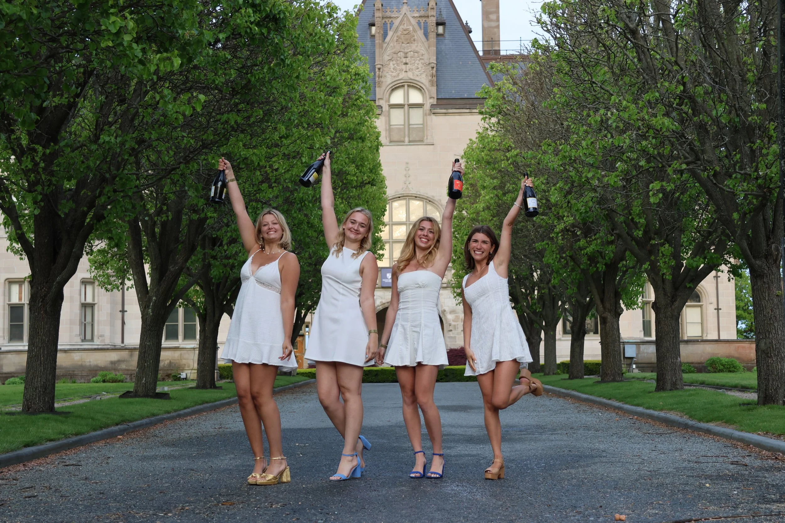 Four women in white dresses standing on a paved road surrounded by green trees, each holding a bottle, with a historic building in the background.