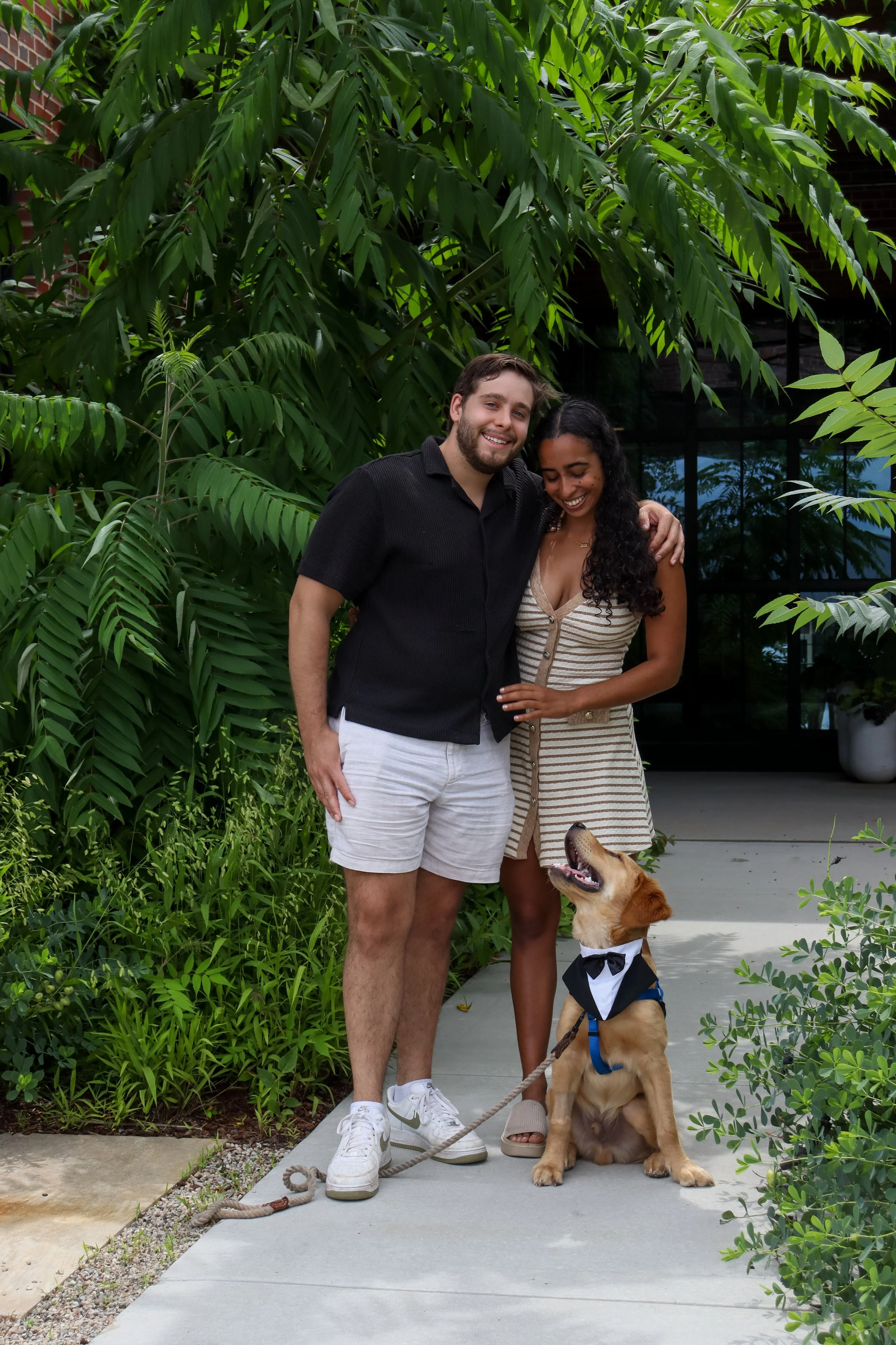 A young couple smiling and hugging outside, with a golden retriever sitting between them wearing a tuxedo and bow tie, on a sidewalk surrounded by lush green plants.