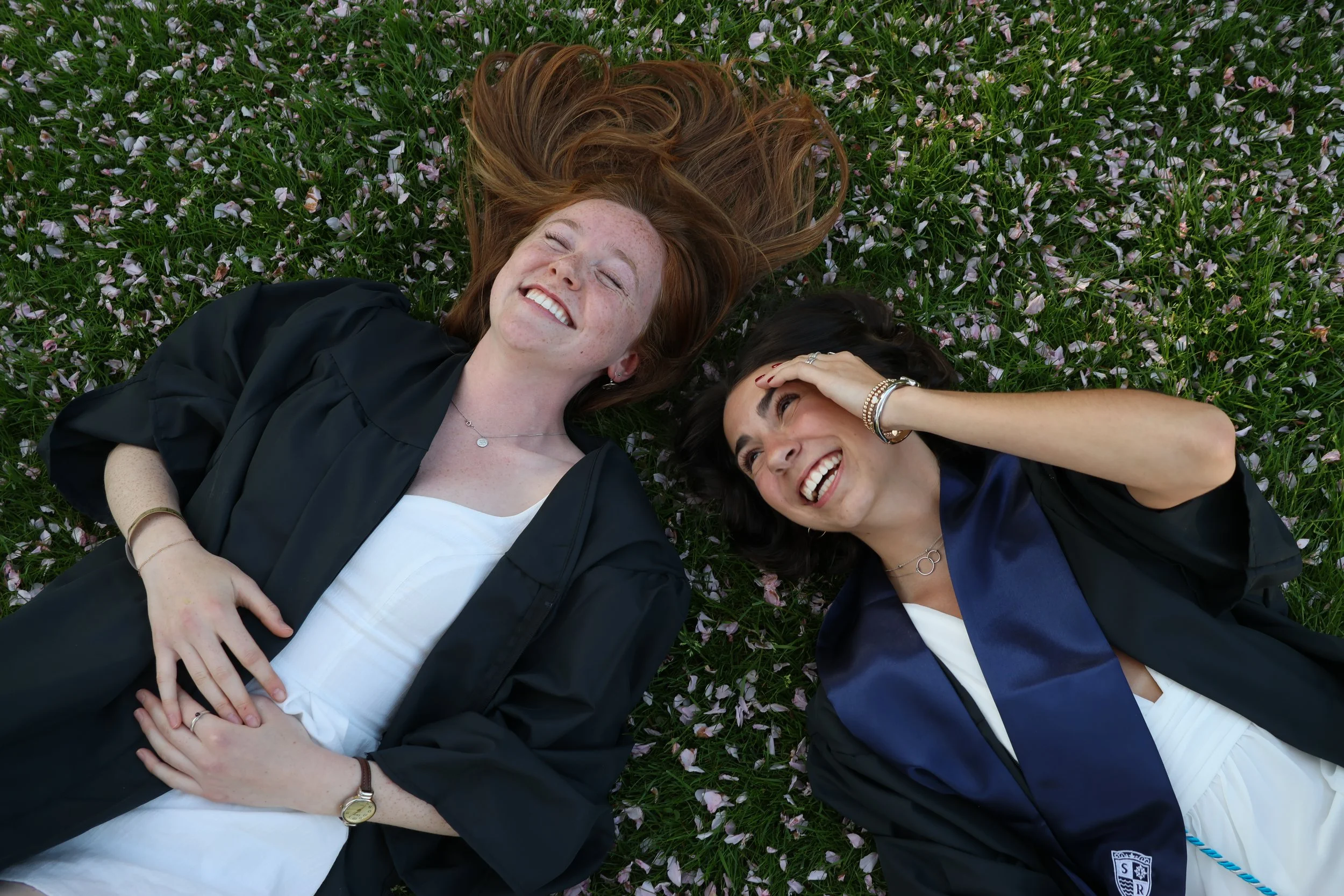 Two young women lying on grass covered with pink flower petals, wearing graduation gowns and smiling.