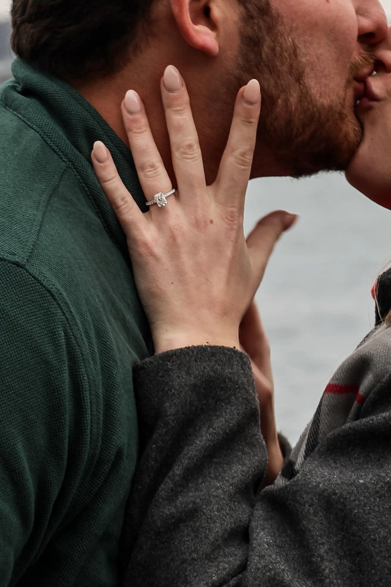 Close-up of a woman showing her engagement ring, touching a man's neck and shoulder as they appear to be about to kiss.