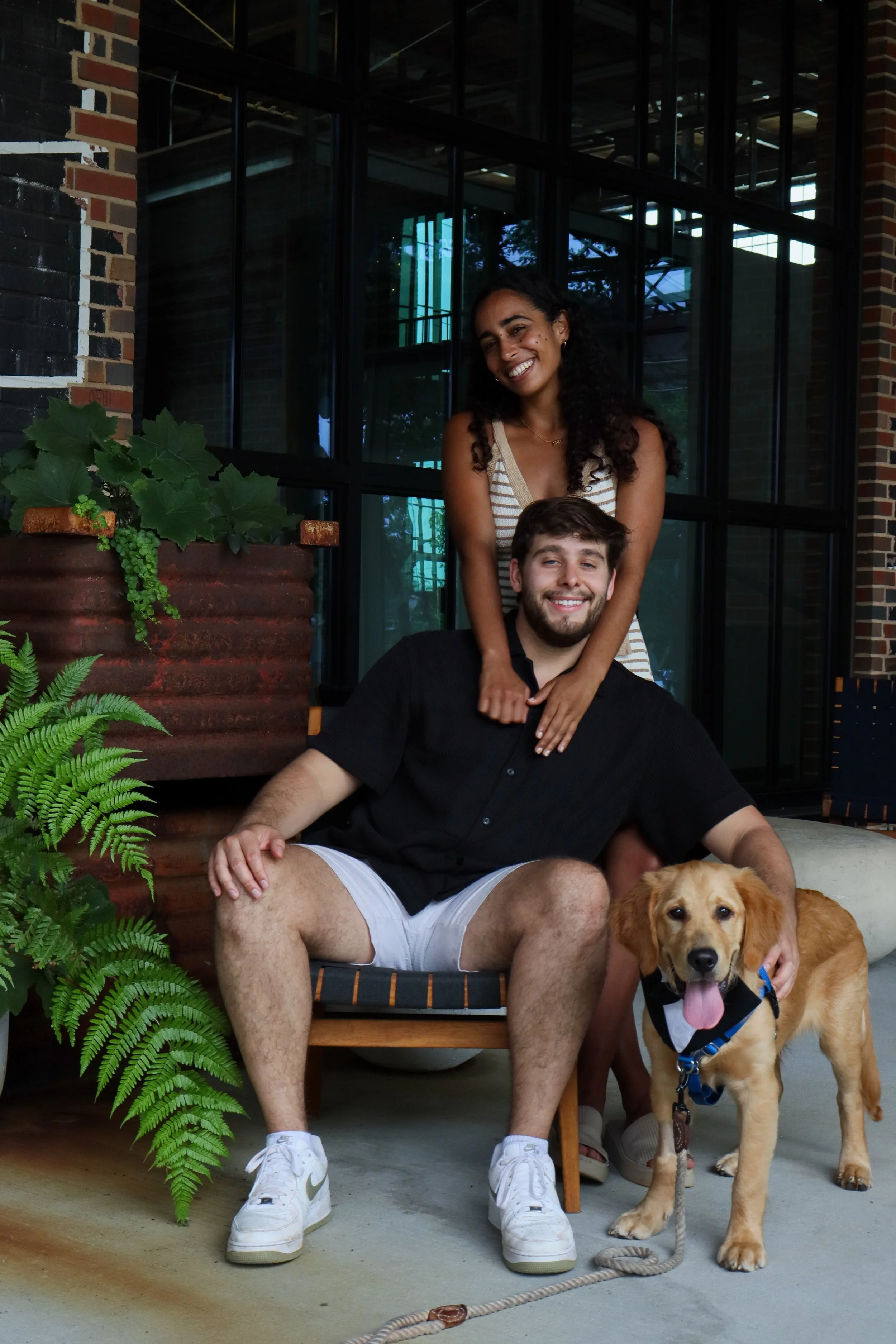 A smiling young woman and man pose with a Golden Retriever dog in front of a large window and brick wall, with green plants nearby.