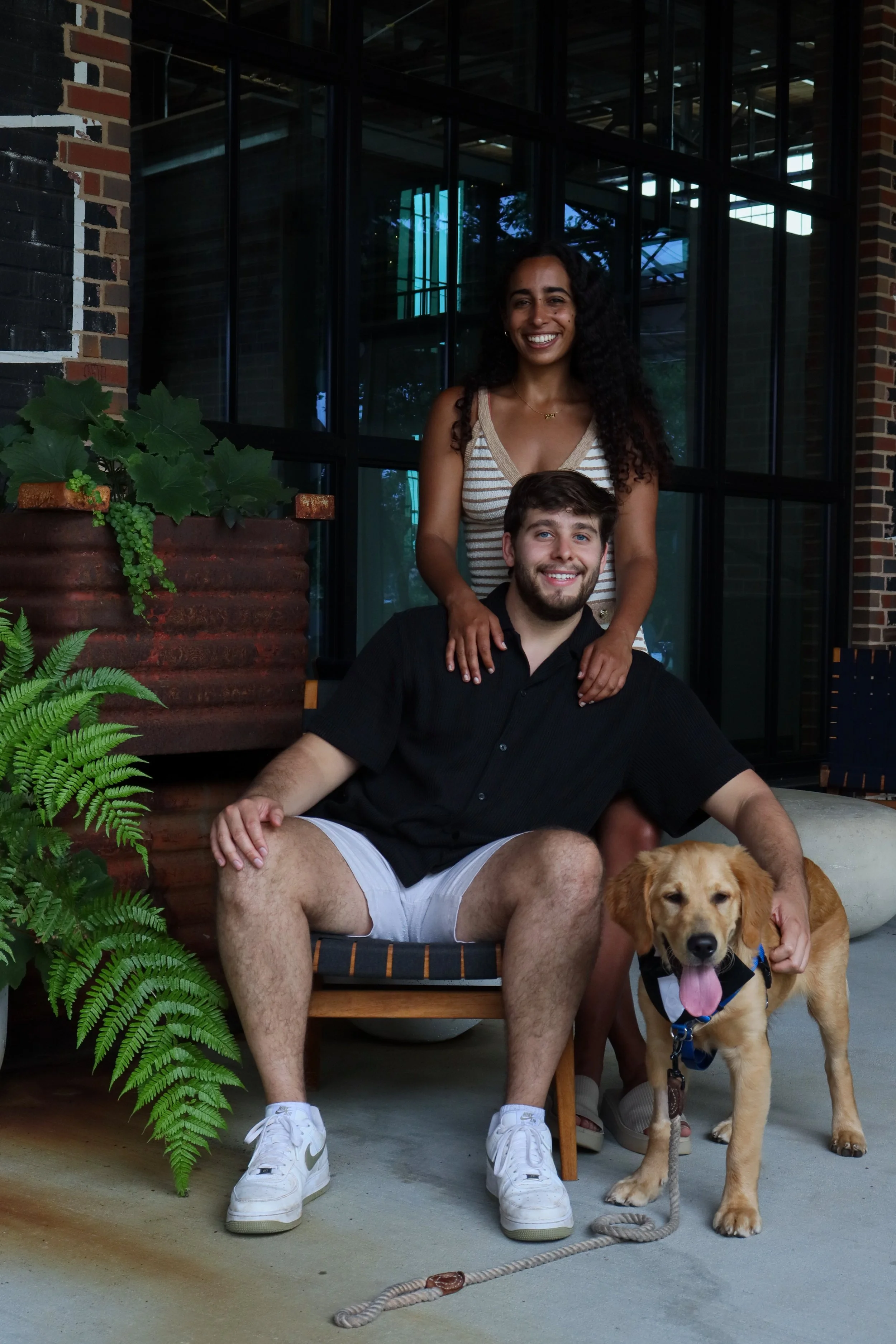 A happy couple, a woman with curly hair and a man with a beard, poses with their dog outside a modern building with large glass windows, surrounded by potted plants.