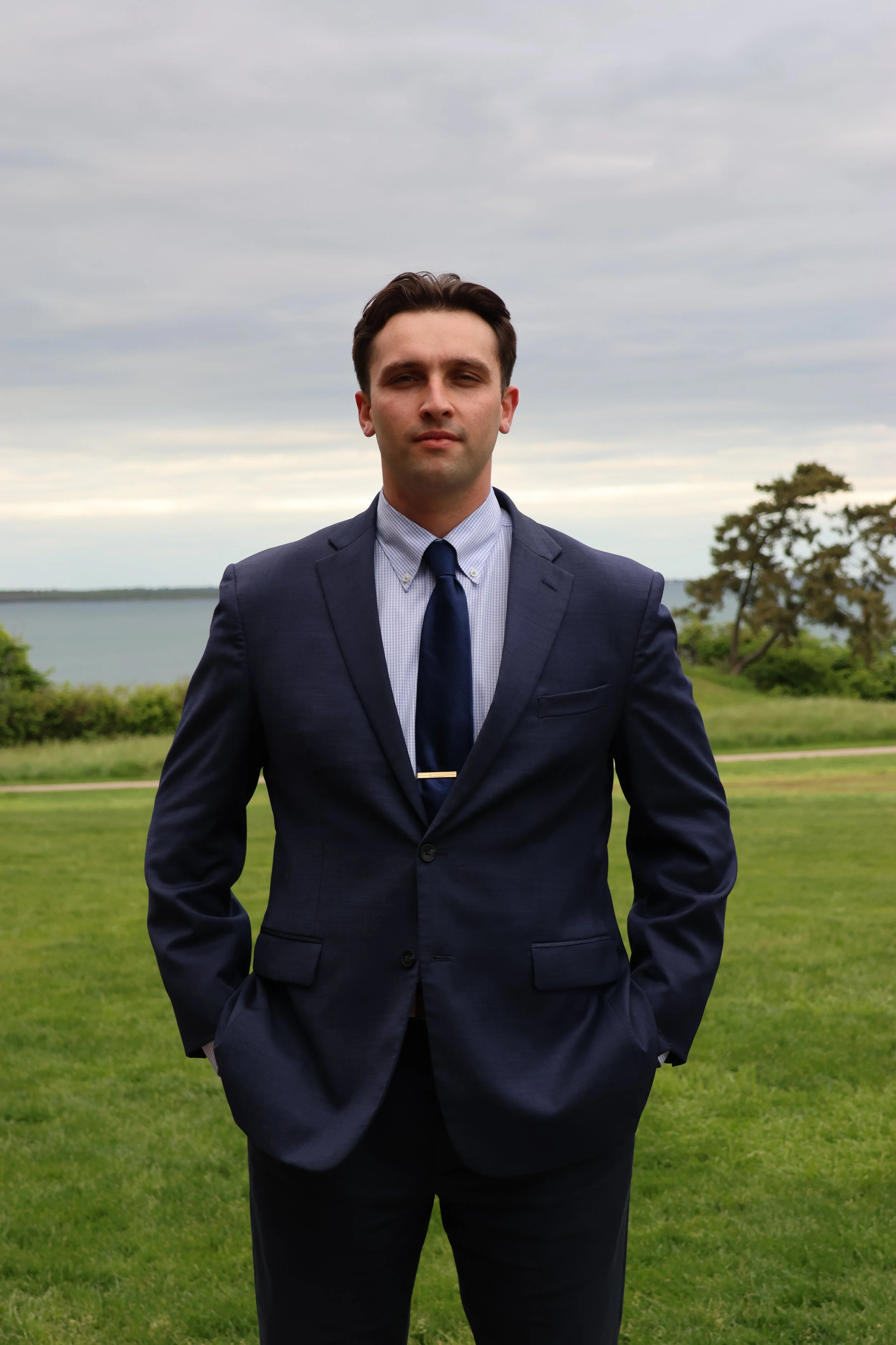 A young man in a navy blue suit and tie stands outdoors with his hands in his pockets. Behind him is a grassy area, trees, and a body of water under a cloudy sky.