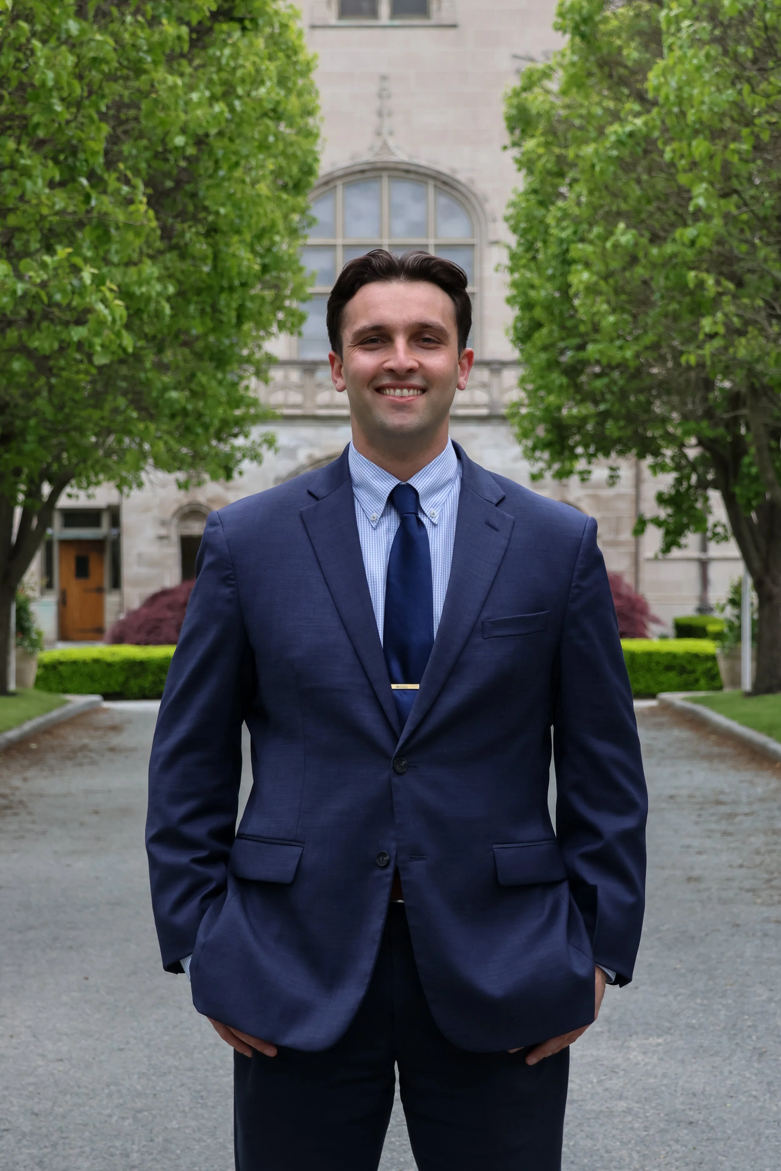 A young man in a navy suit, light blue dress shirt, and matching tie standing on a paved pathway in front of a historic building with large windows, surrounded by green trees and manicured bushes, smiling at the camera.
