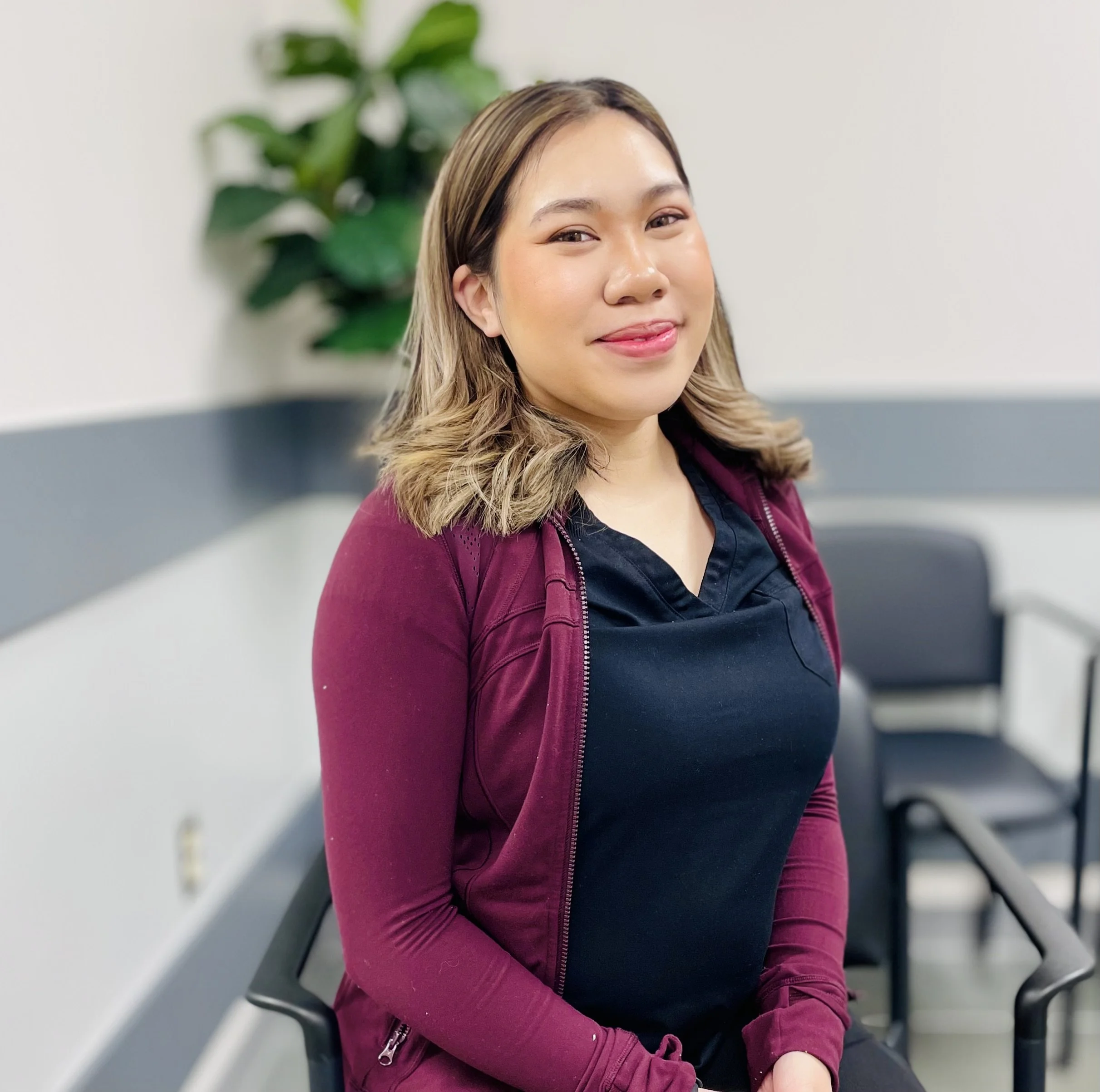 A young woman with shoulder-length light brown hair, wearing a black top and a burgundy jacket, sitting in a waiting area with black chairs and a green plant in the background.