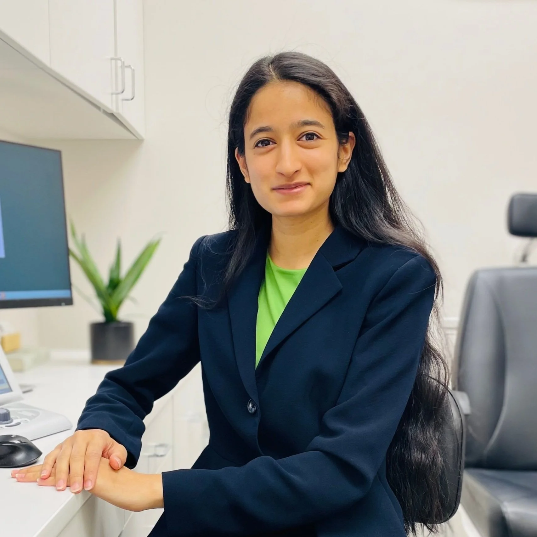 A woman with long dark hair, wearing a black blazer over a bright green shirt, sitting at a desk in an office with a computer, a potted plant, and a monitor in the background.