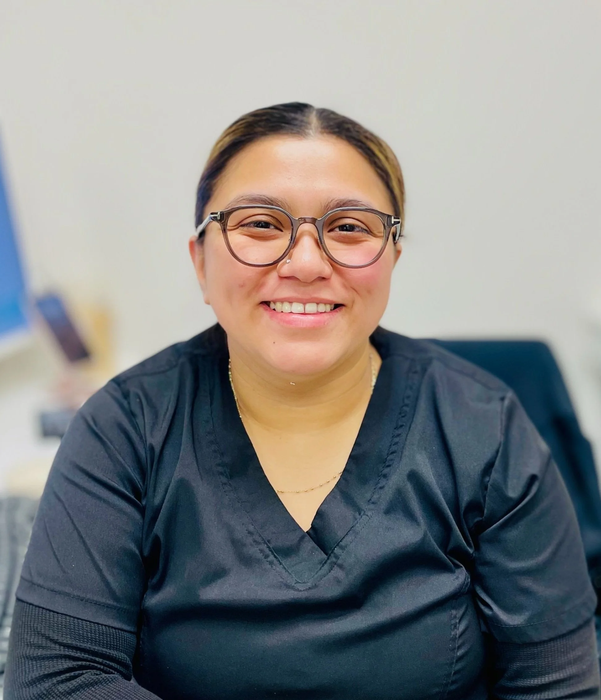 A woman wearing glasses and a black medical scrubs, sitting in an office or clinic, smiling at the camera.