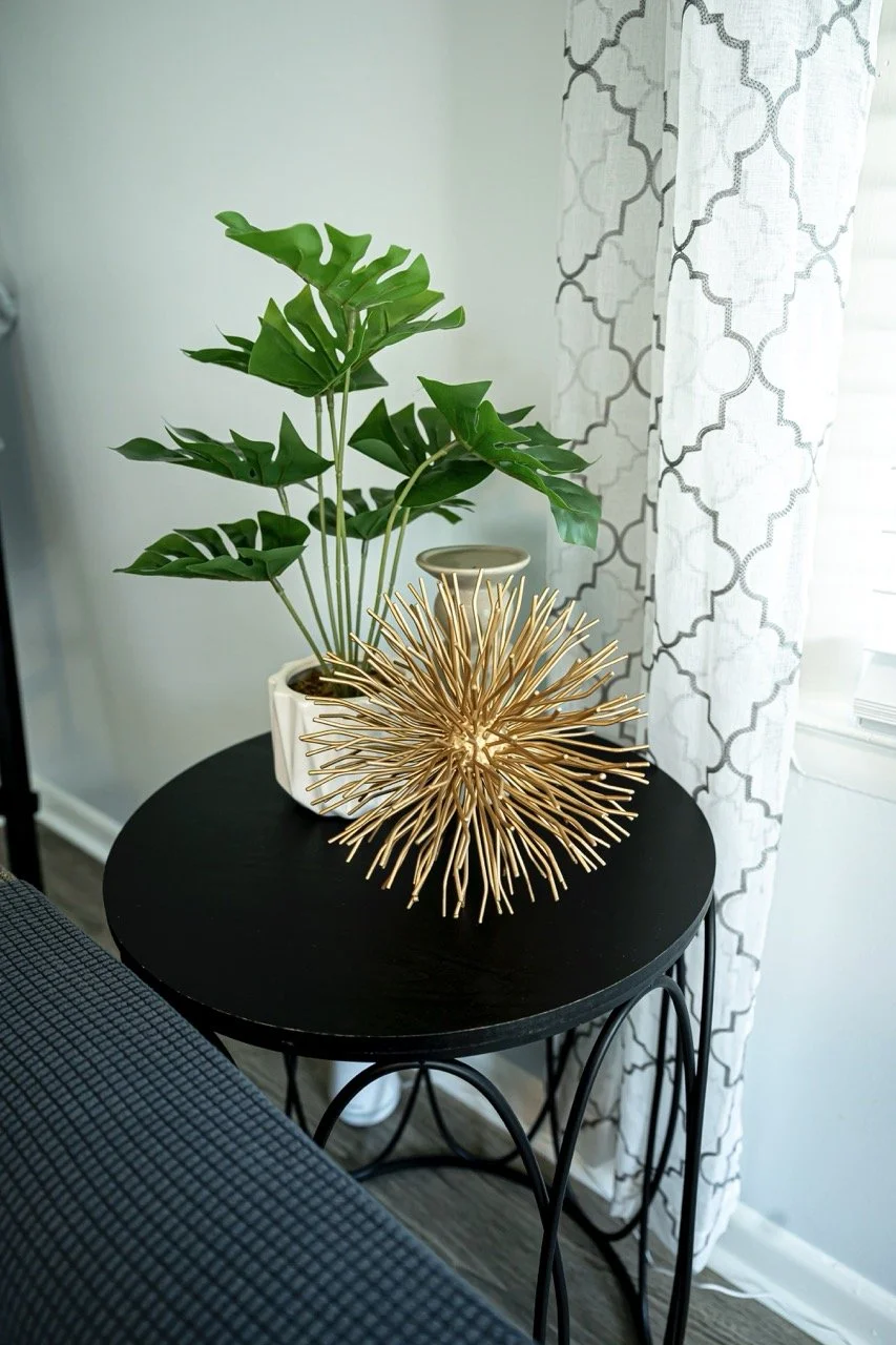 A round black side table with a white pot containing a green monstera plant, a small empty white bowl, and a gold decorative spiky sphere, situated near a window with white patterned curtains.