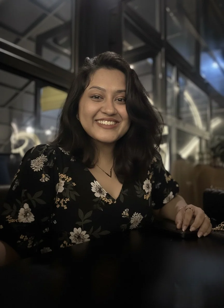 A woman with dark, wavy hair smiles at the camera, sitting at a table in a dimly lit restaurant or cafe, wearing a black floral dress with white and beige flowers.