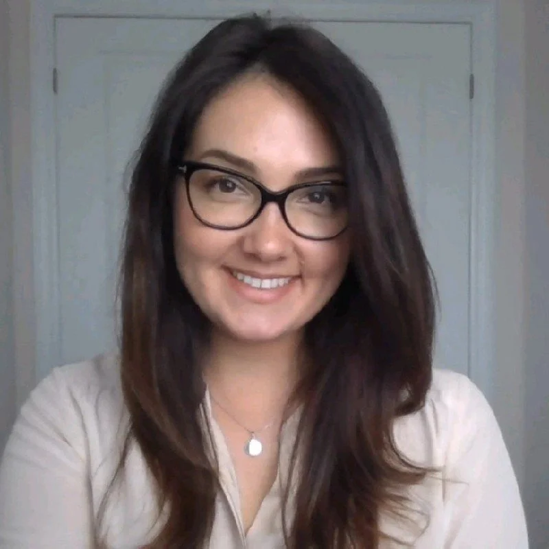 A woman with long dark hair, wearing glasses and a light-colored shirt, smiling at the camera.
