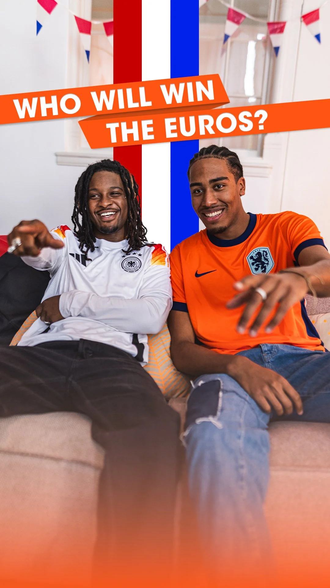 Two young men sitting on a beige couch, wearing soccer jerseys of Germany and the Netherlands, in a living room decorated with red, white, and blue bunting.