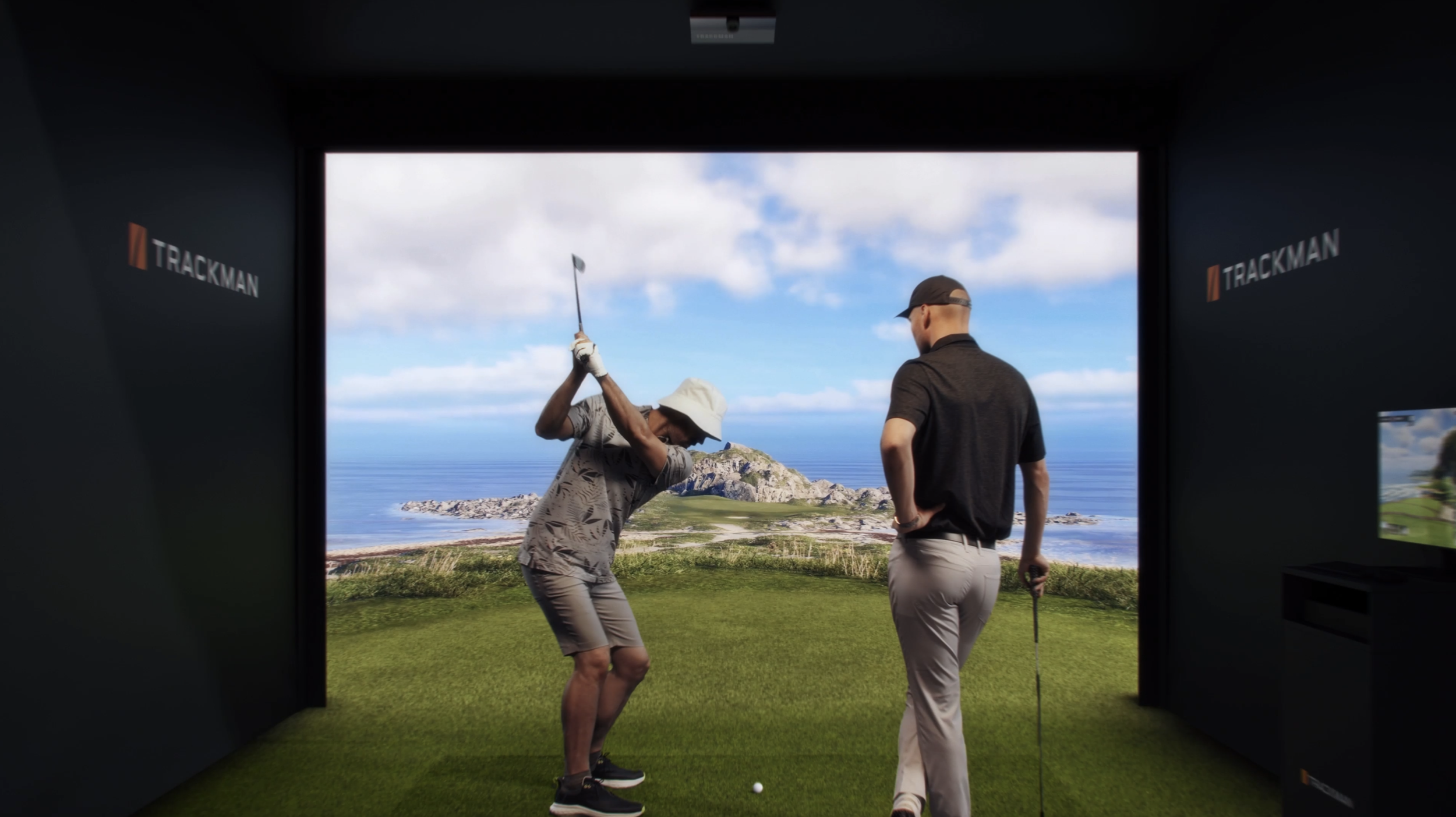 Two men are in a golf simulation room, one preparing to hit a golf ball with a club, while the other watches. The room has a large screen showing a scenic outdoor golf course with ocean and hills in the background.