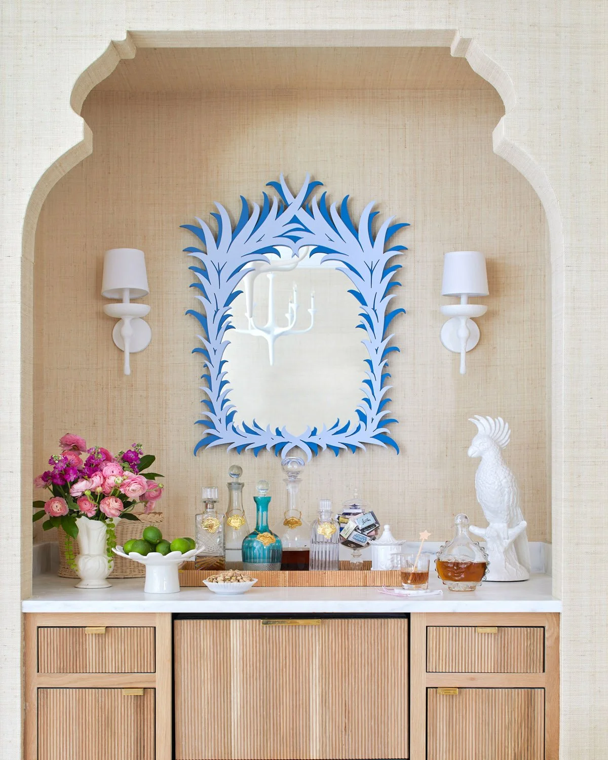 Decorative bar or sideboard area with a large blue-framed mirror, white wall sconces, a vase of pink flowers, green apples, various bottles, and a white ceramic bird sculpture