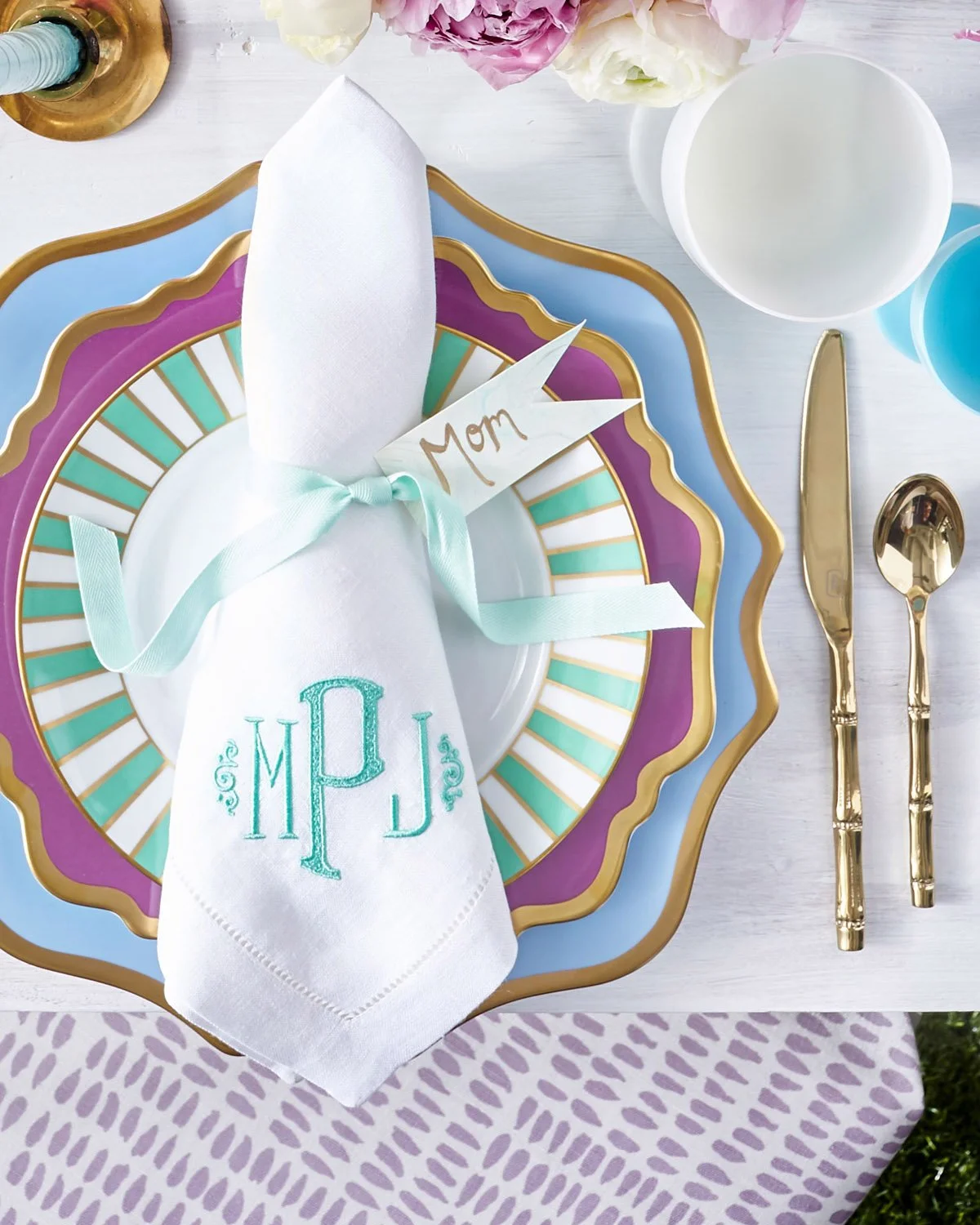 A folded mongram napkin with a placetag labeled "mom" on top of a blue and purple table setting
