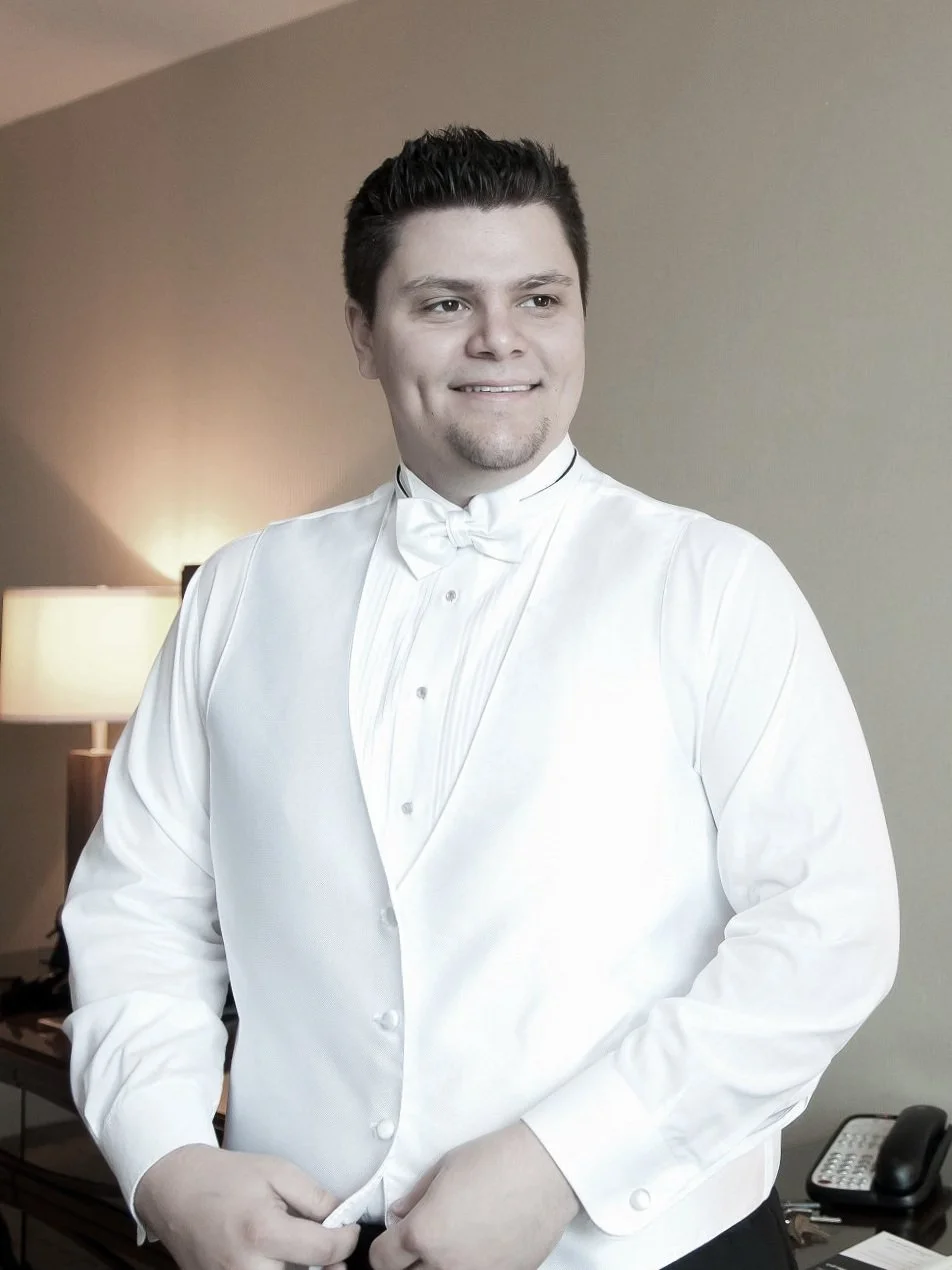 A man dressed in a white tuxedo with a bow tie standing in a room with a lamp and phone on a desk.