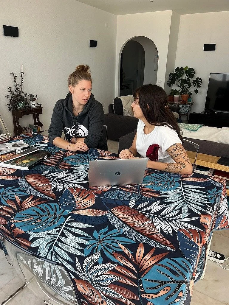 Two women sitting at a table with a leaf-patterned tablecloth, having a conversation. One is using a laptop, and there are plants and a TV in the background.