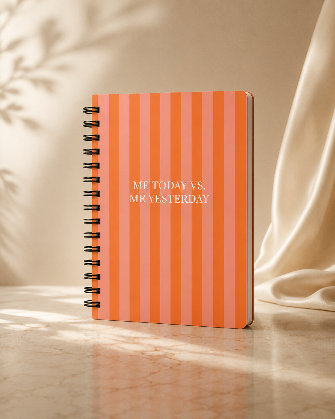 Spiral-bound notebook with orange and pink vertical stripes on the cover and white text that reads 'ME TODAY VS. ME YESTERDAY' placed on a marble surface with a beige fabric backdrop.
