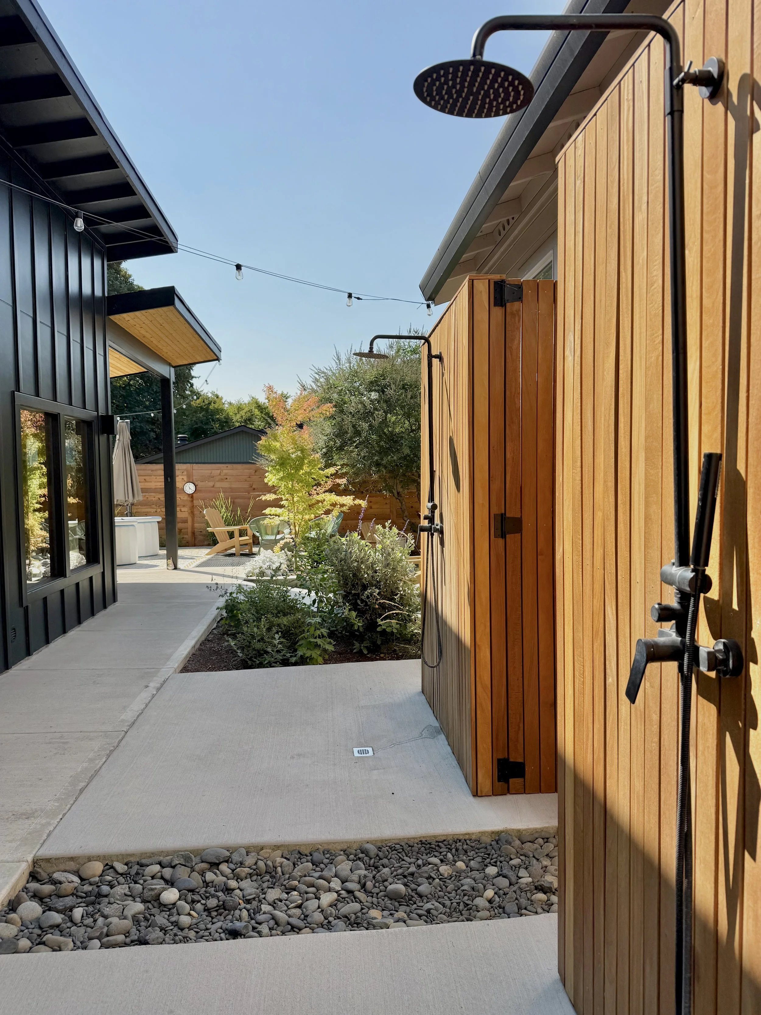 Outdoor shower with wooden framing in a backyard, with a patio and garden area, string lights overhead, and neighboring houses in the background.