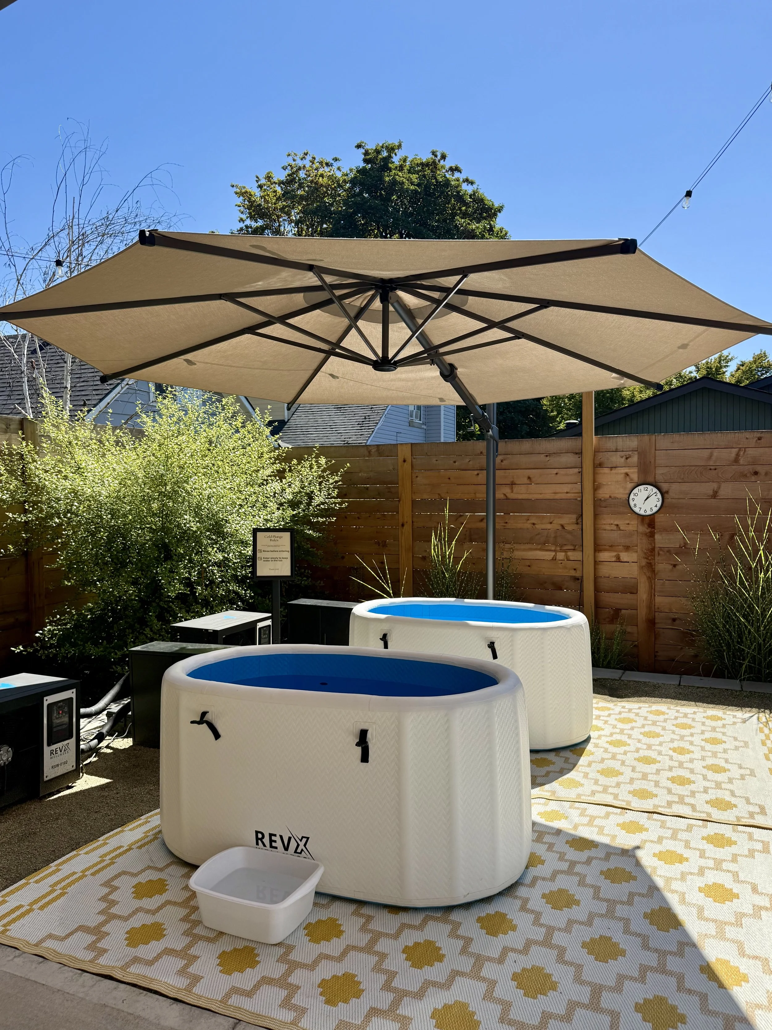 Backyard patio with two white inflatable hot tubs, wooden outdoor chairs, purple flowering bush, wooden privacy fence, string lights, and a partly cloudy blue sky.