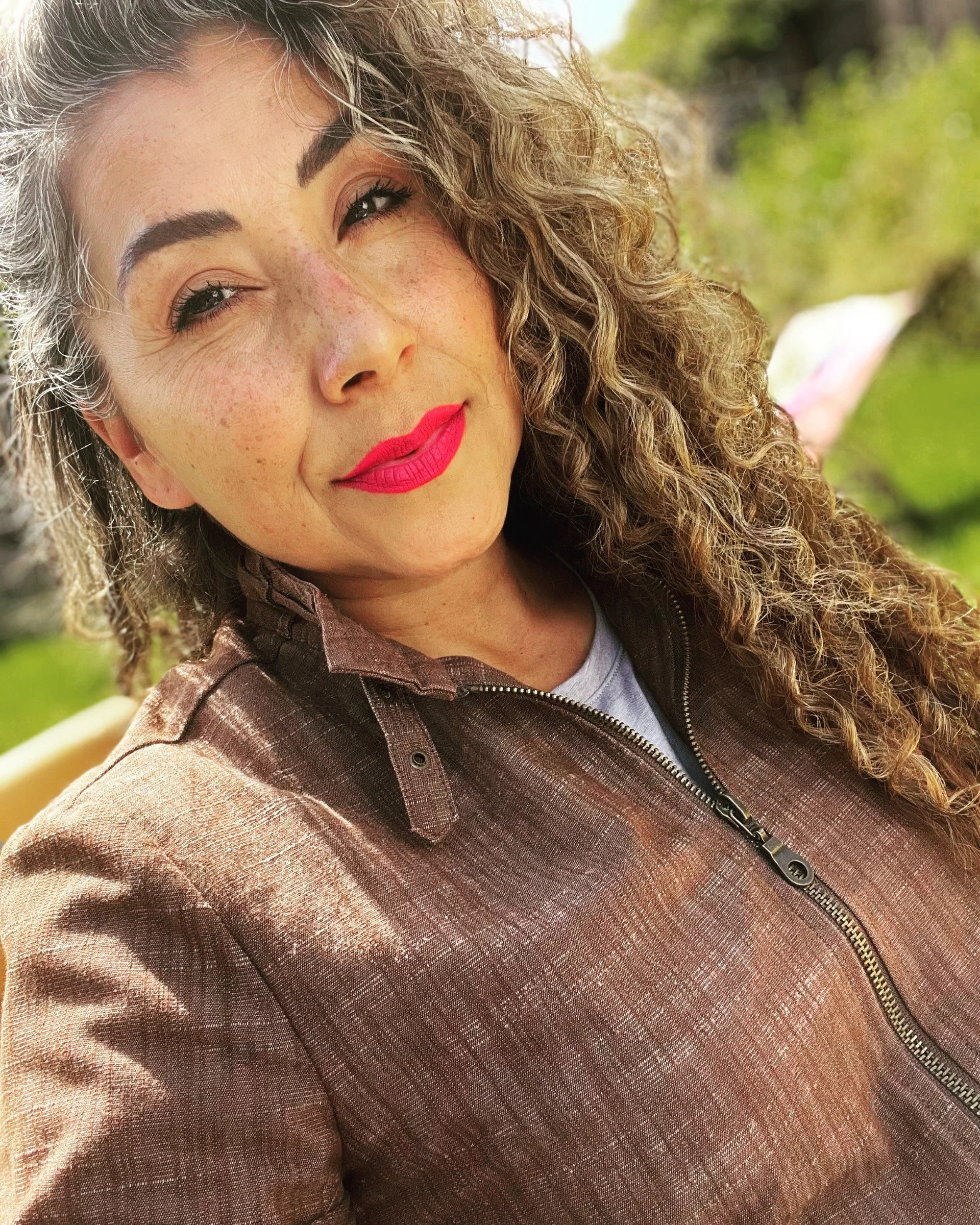 A woman with curly, gray and black hair smiling at the camera, wearing makeup with purple lipstick, in front of a blurred, light-colored background.