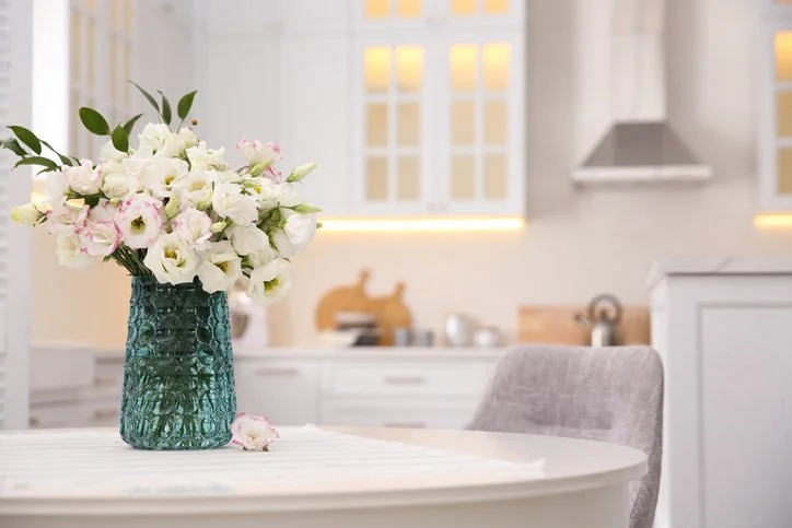 A bouquet of white and pink flowers in a textured green vase on a white table in a bright, modern kitchen with white cabinets and a window.
