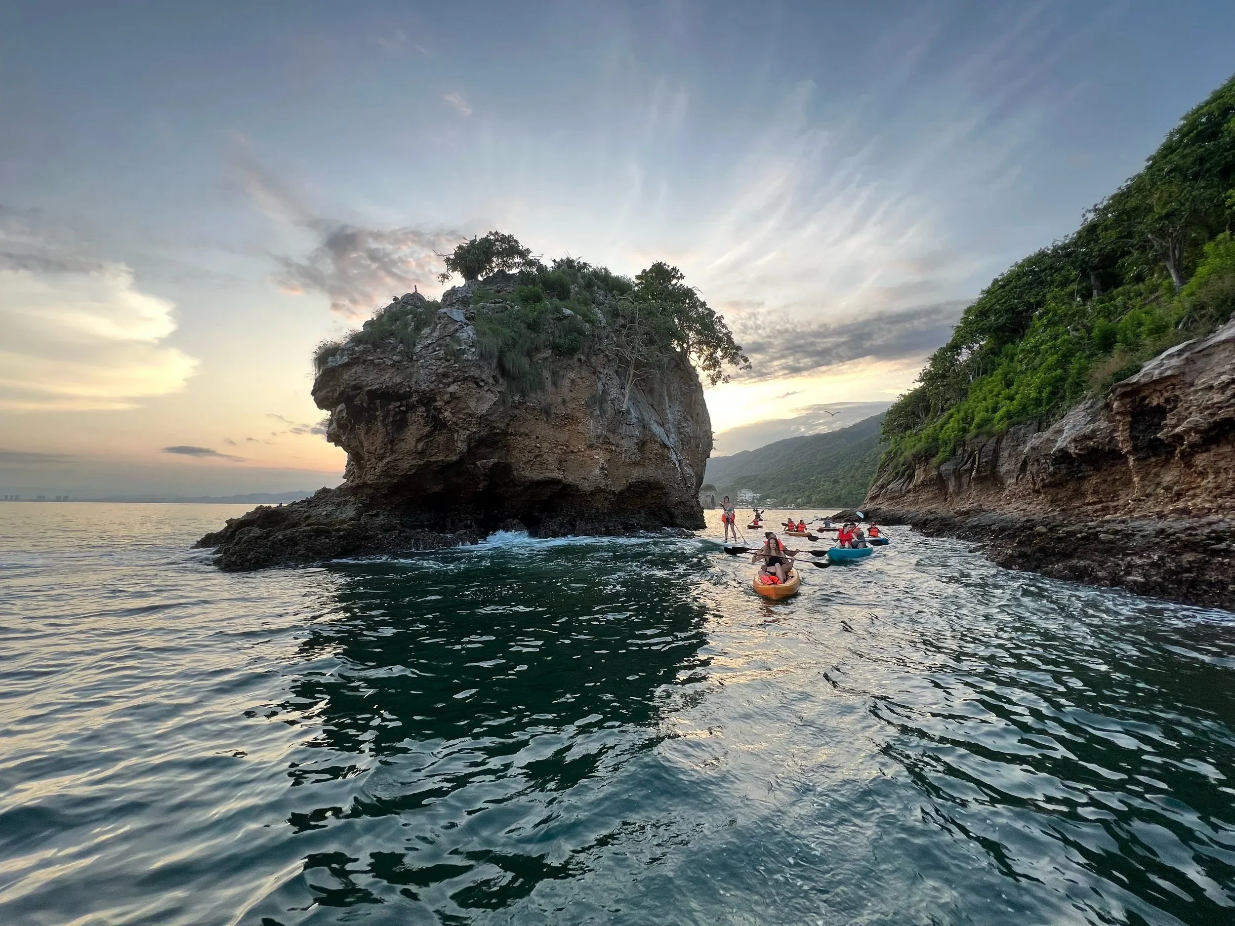 People kayaking near a large rock formation on the ocean at sunset with hillside covered in greenery.