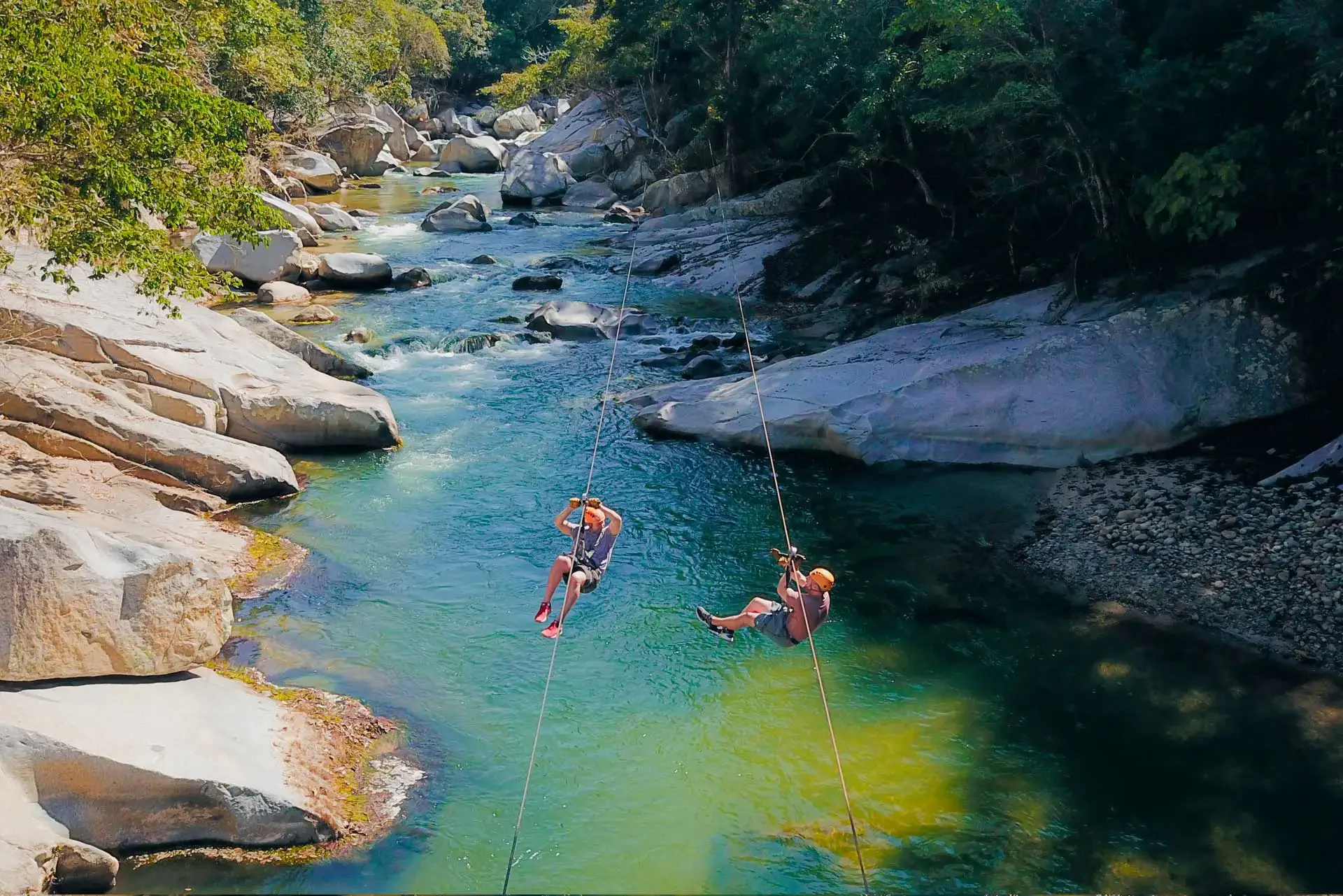 Two children wearing helmets and harnesses are ziplining over a river with clear blue-green water, surrounded by large rocks and green trees.