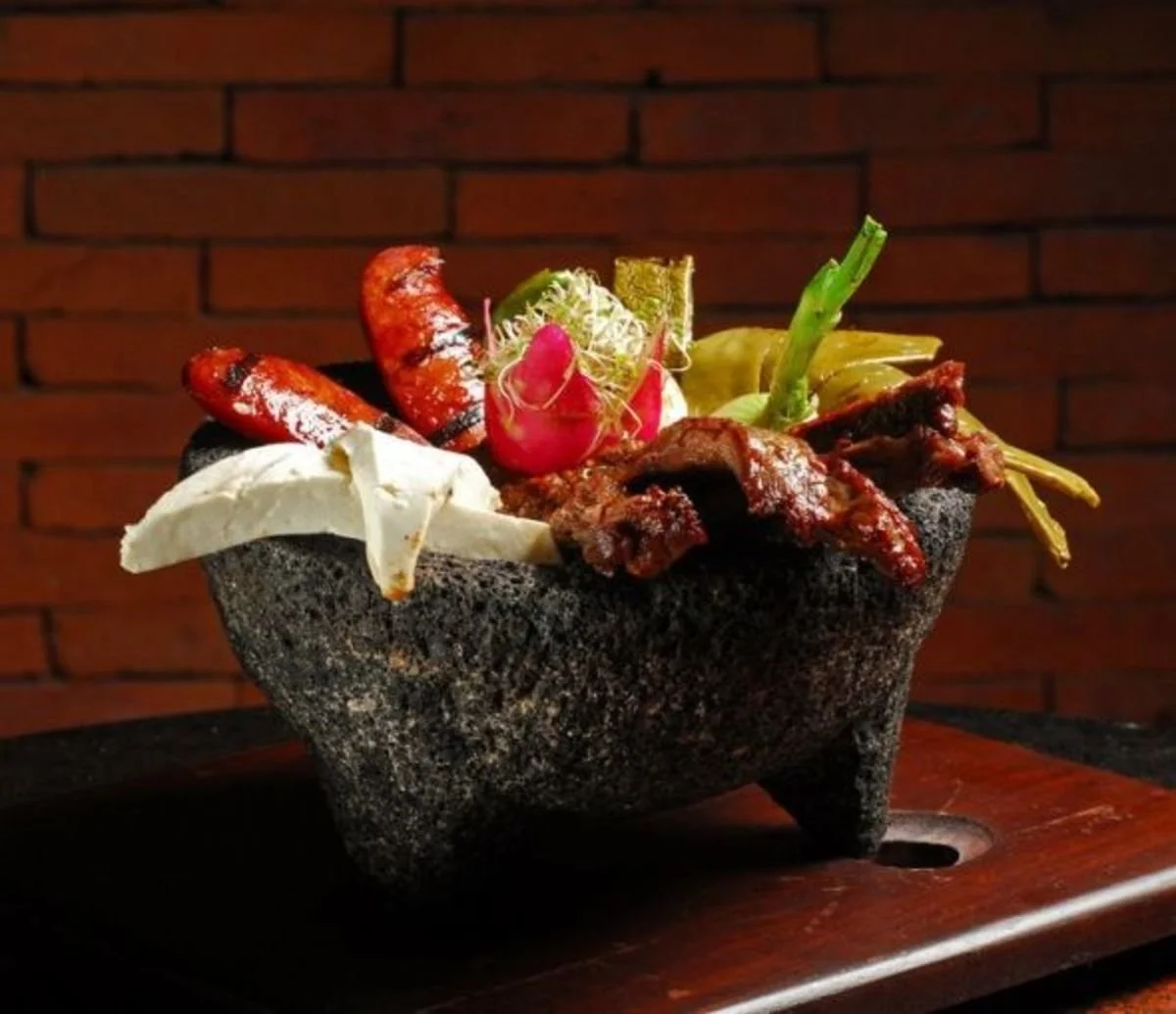 A stone molcajete filled with grilled meats, vegetables, and cheese, set against a brick wall background.